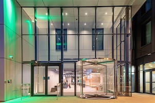 Una entrada de edificio moderna con puertas de vidrio y un giratorio, que da acceso a un interior con zona de estar y una escalera.