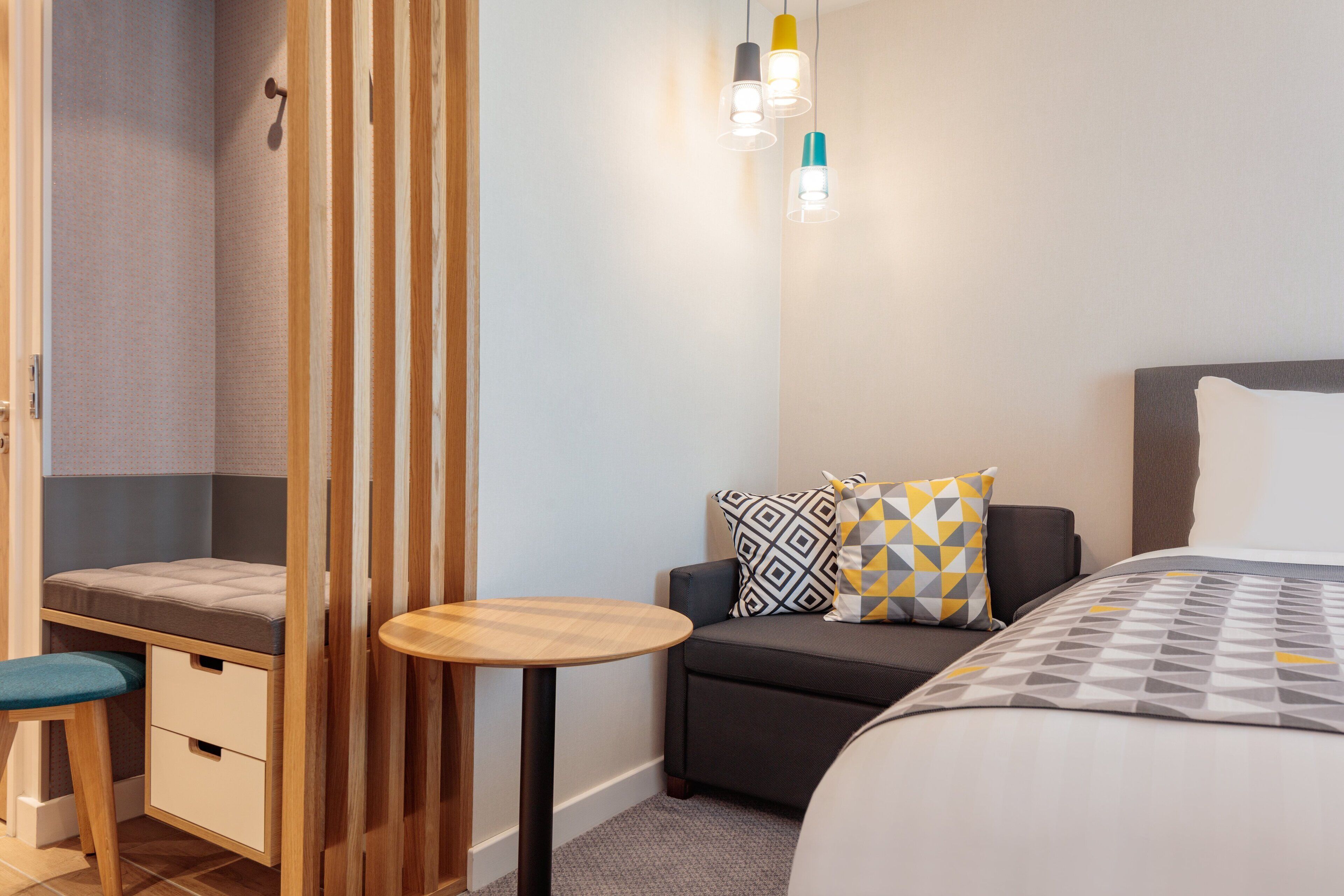 Habitación de hotel compacta con una cama, un pequeño rincón de descanso, un separador de madera y una mesa redonda.
