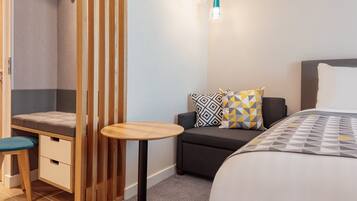 Habitación de hotel compacta con una cama, un pequeño rincón de descanso, un separador de madera y una mesa redonda.