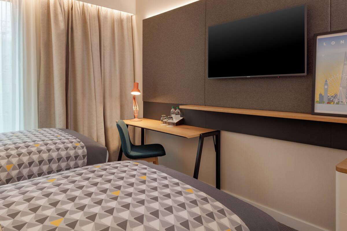 A hotel room with two beds, a desk, a chair, a TV, and a framed picture on the wall.