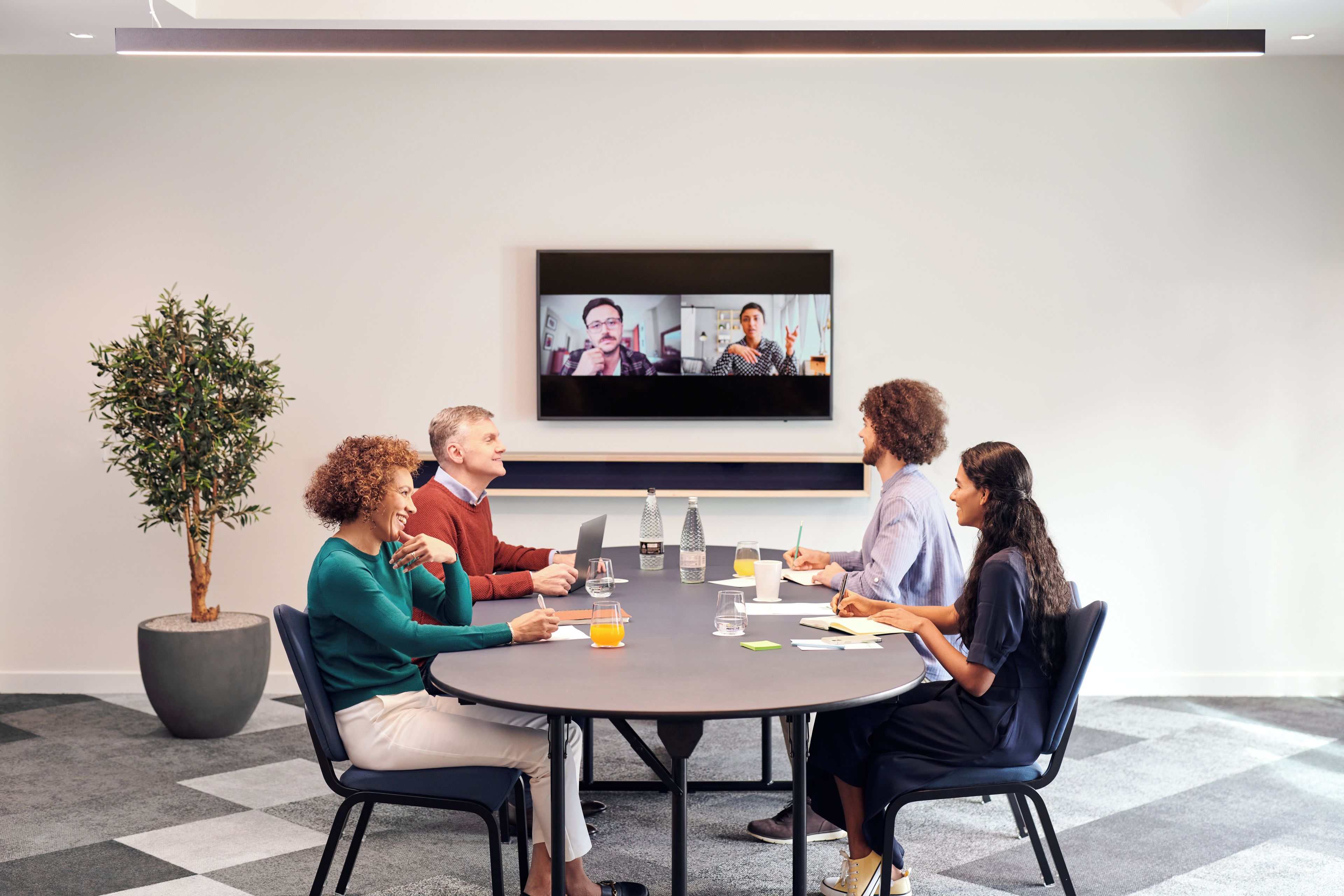 A conference room with a round table, chairs, a television screen, and a potted plant.