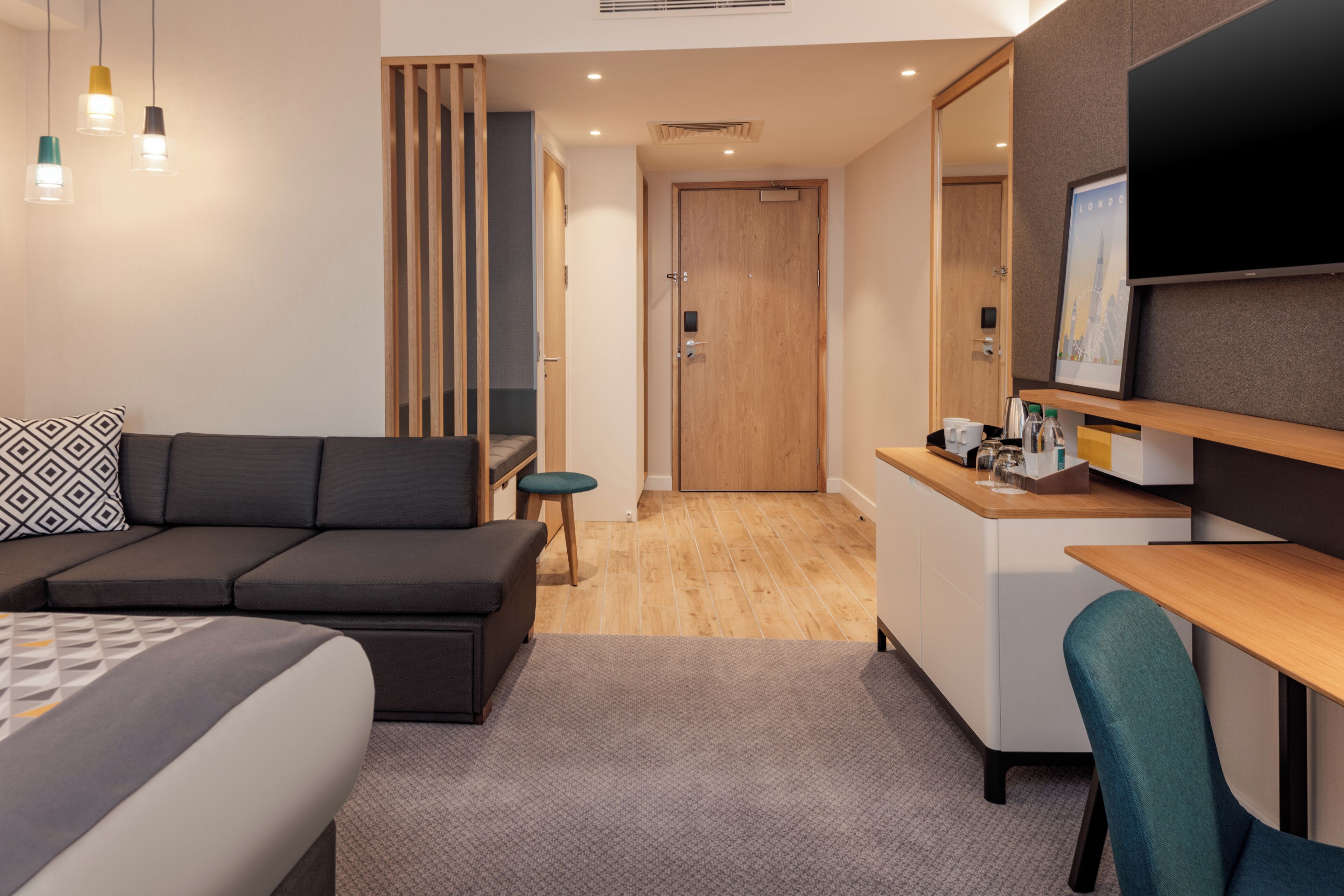A modern hotel room with a sofa, a desk, a chair, a bed, a TV, and a door leading to a bathroom.