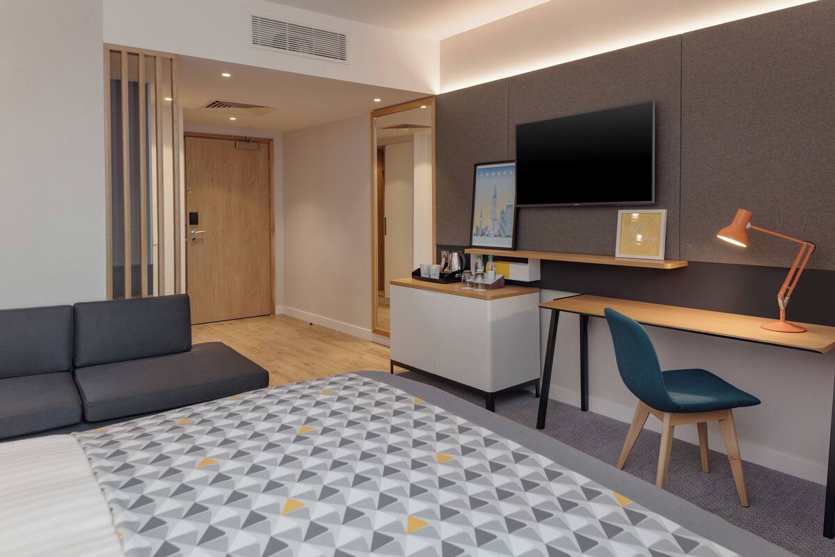 A modern hotel room with a bed, a desk, a chair, a TV, and a sofa.
