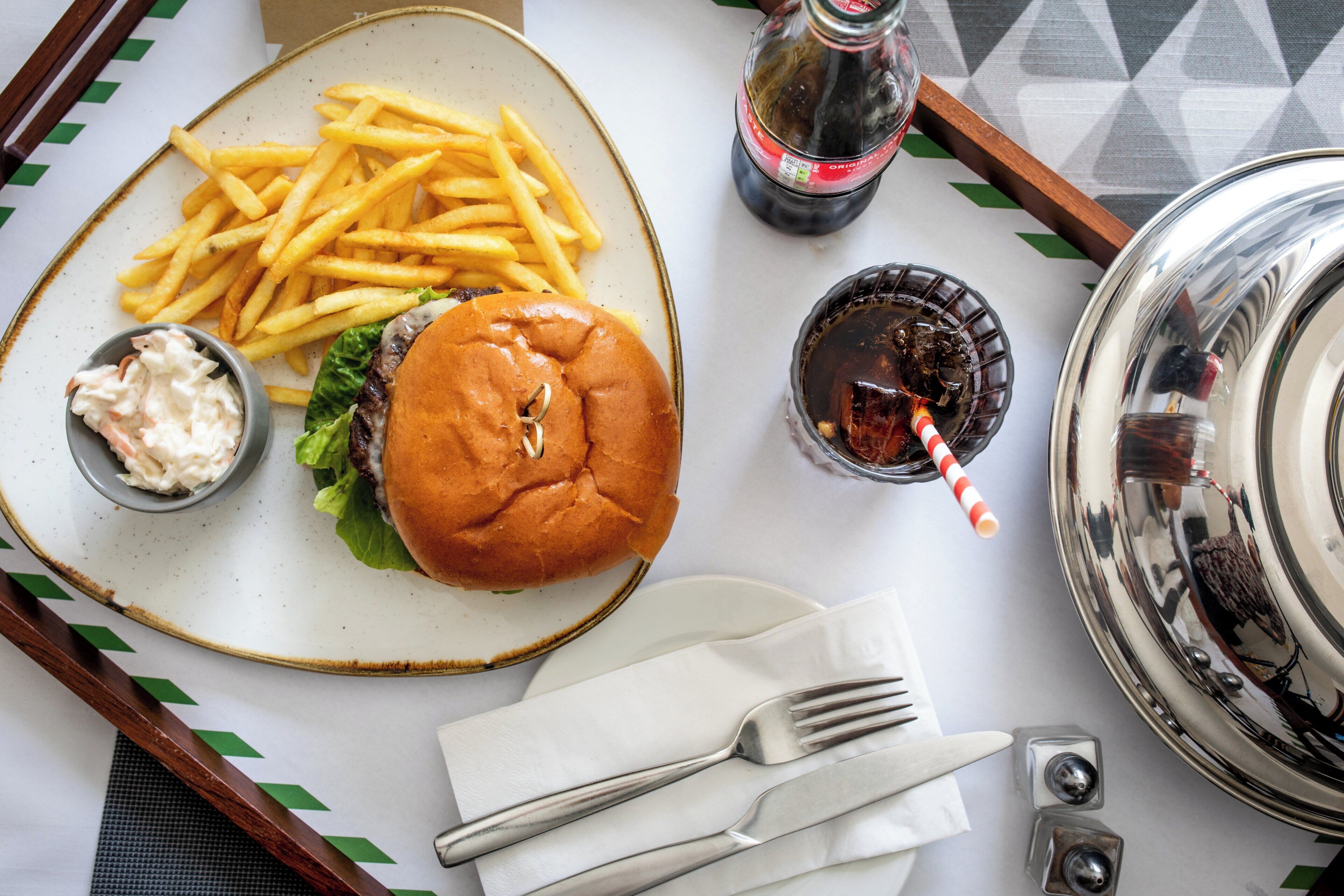 A meal consisting of a burger, fries, and coleslaw on a plate, accompanied by a soda and a glass with a straw.