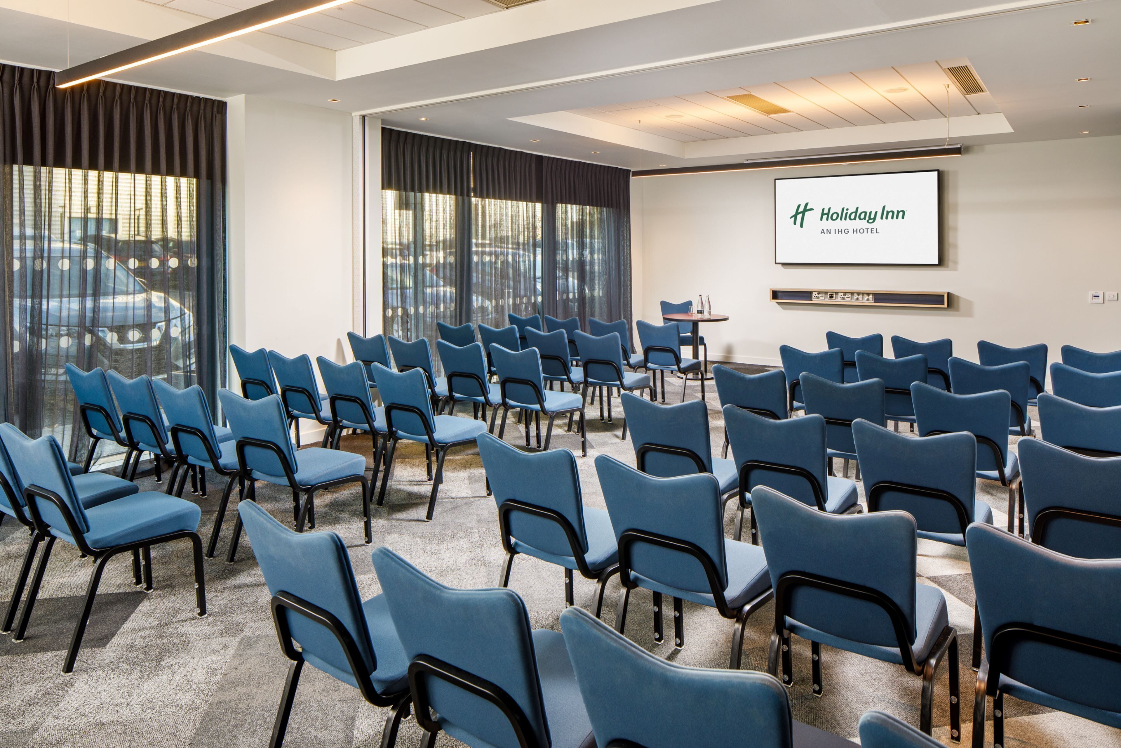 A conference room with rows of blue chairs, a large screen displaying the Holiday Inn logo, and a small table with a projector.
