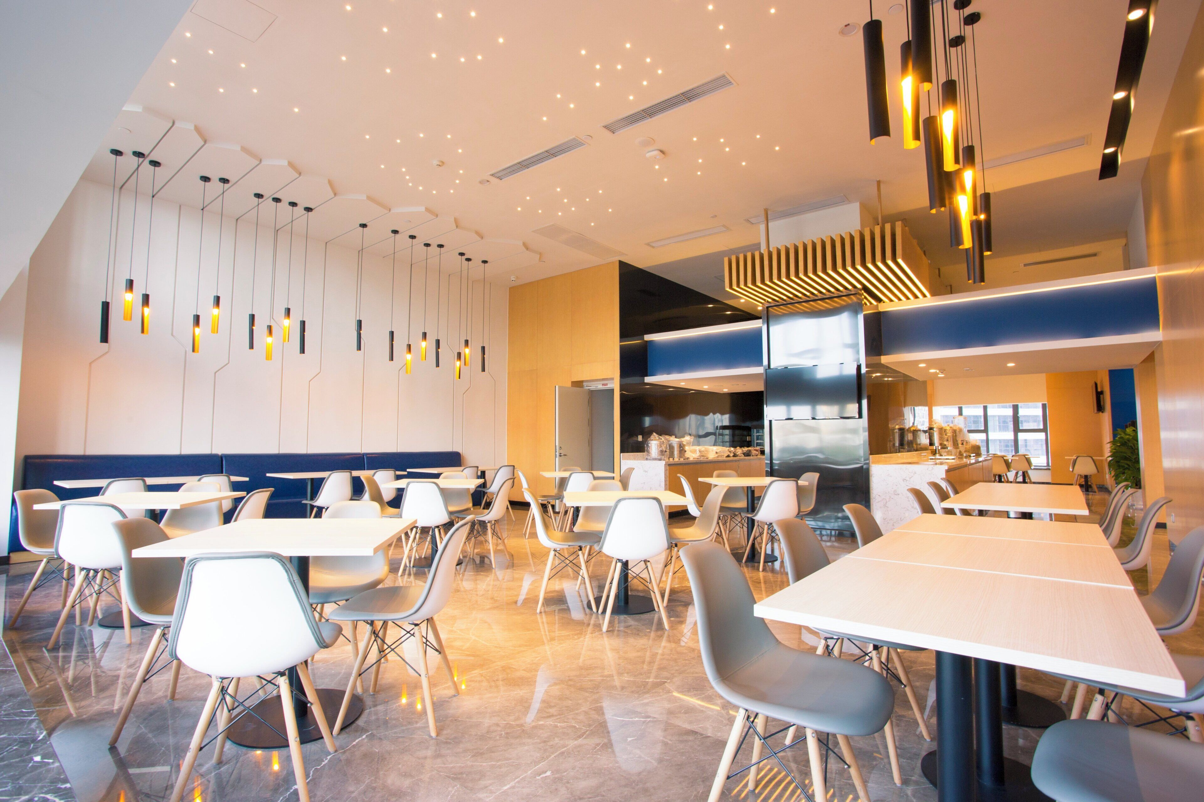 A modern dining area with multiple tables and chairs, a kitchen area, and a ceiling with recessed lighting.