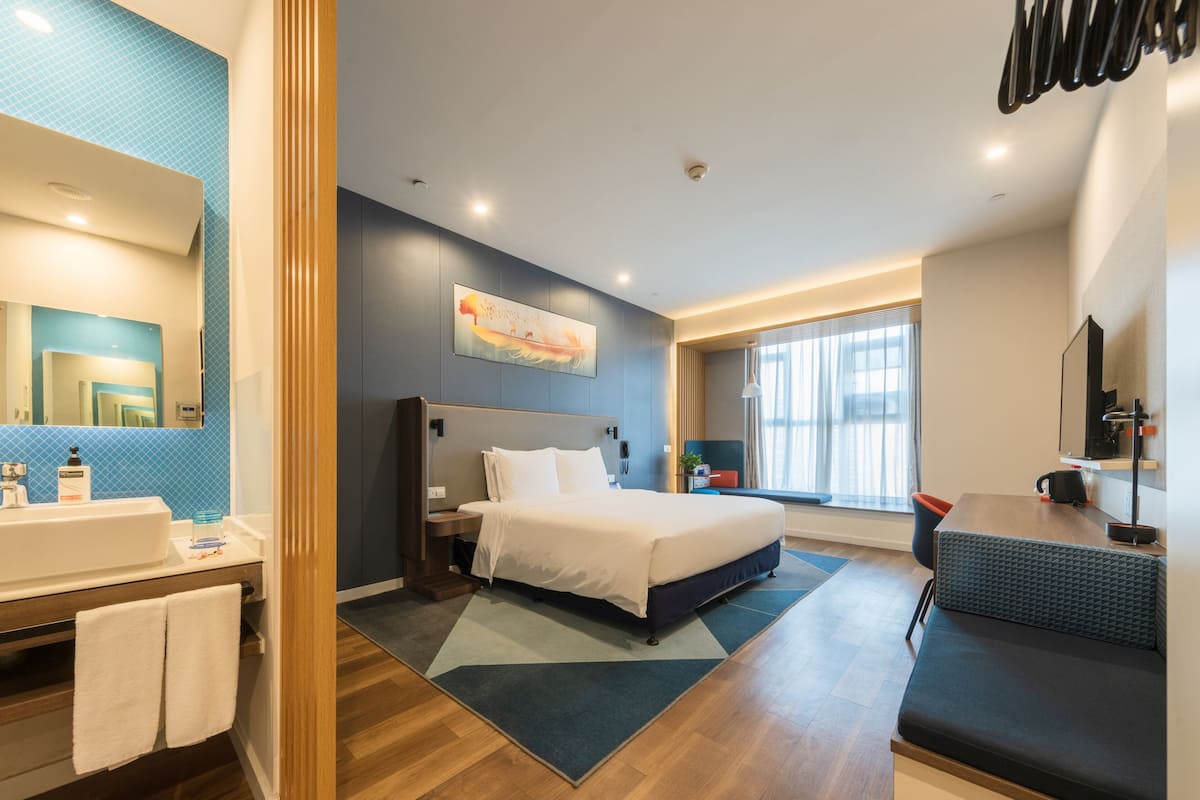 A modern hotel room with a large bed, a bathroom with a sink and mirror, a desk with a chair, and a window with curtains.