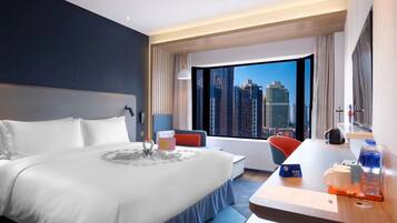 A modern hotel room with a large bed, a desk with a chair, a view of city buildings, and a window with curtains.