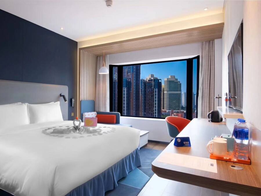 A modern hotel room with a large bed, a desk with a chair, a view of city buildings, and a window with curtains.