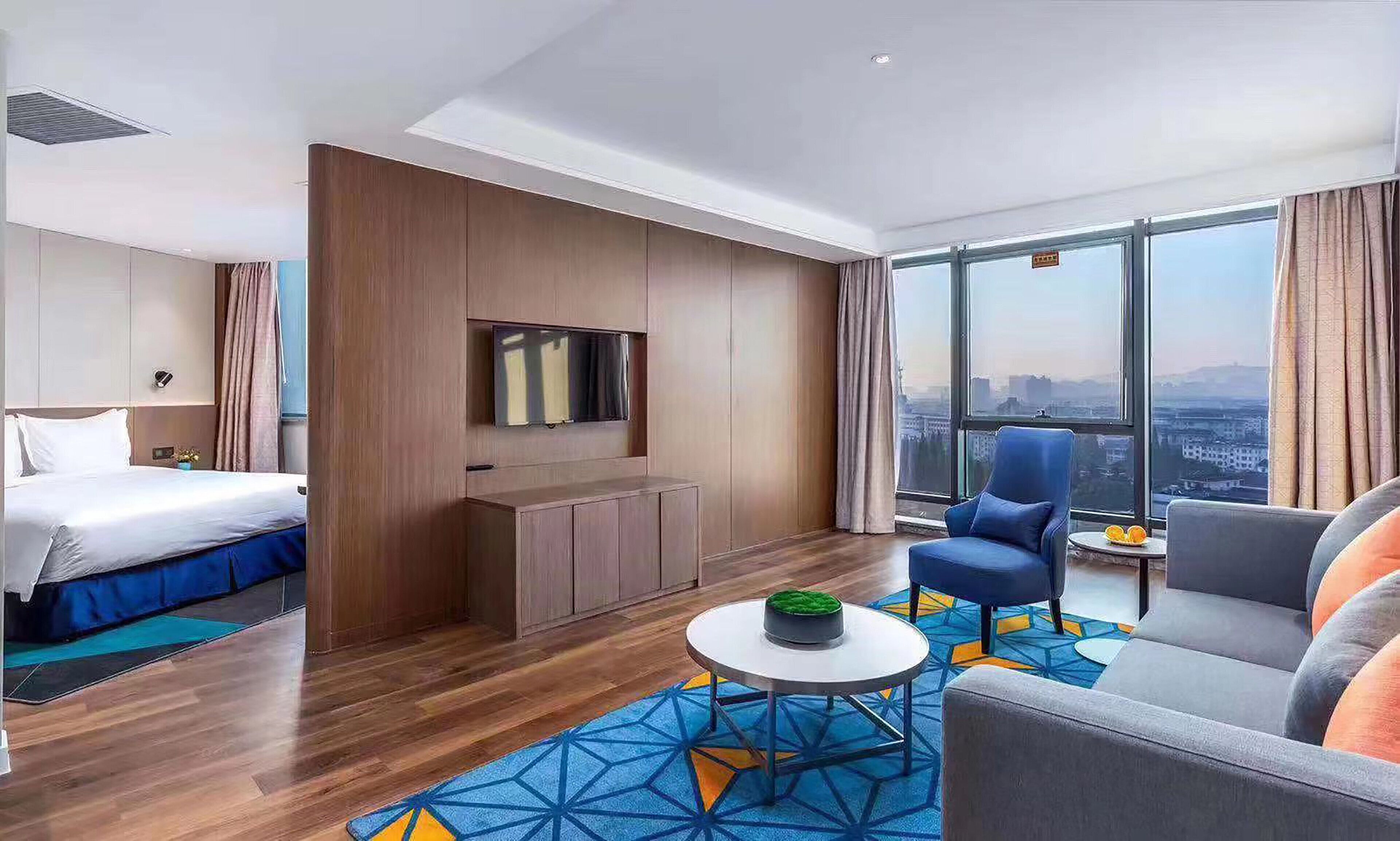 A hotel room with a bed, a seating area with a sofa and armchair, a small table, a TV mounted on a wall, and a large window offering a city view.