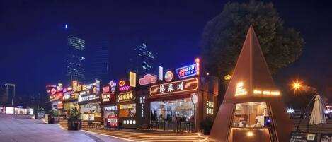 Una calle urbana iluminada de noche, con escaparates luminosos, incluyendo una cafetería con un toldo de forma piramidal distintiva.