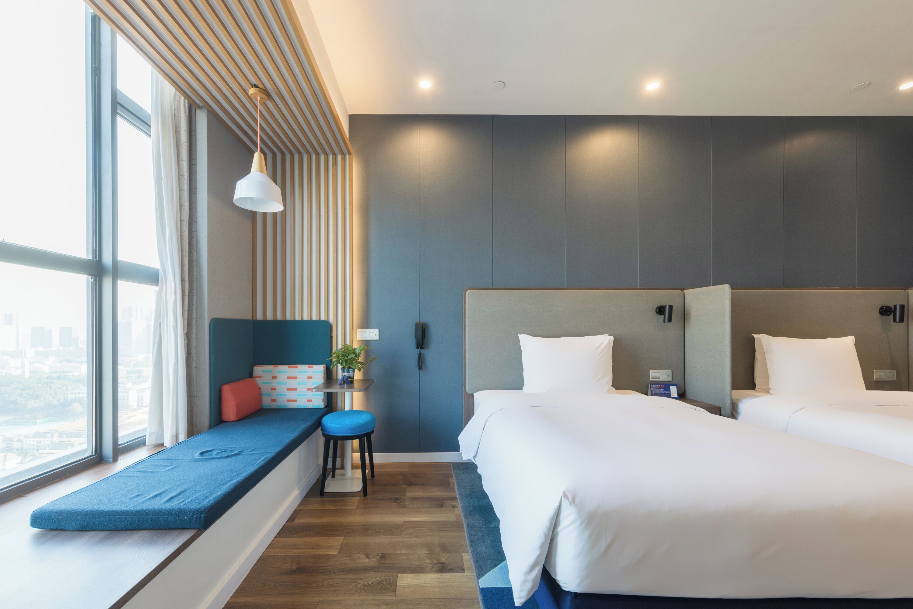A modern hotel room with a large window, a built-in bench, two beds, and a small table with a plant.
