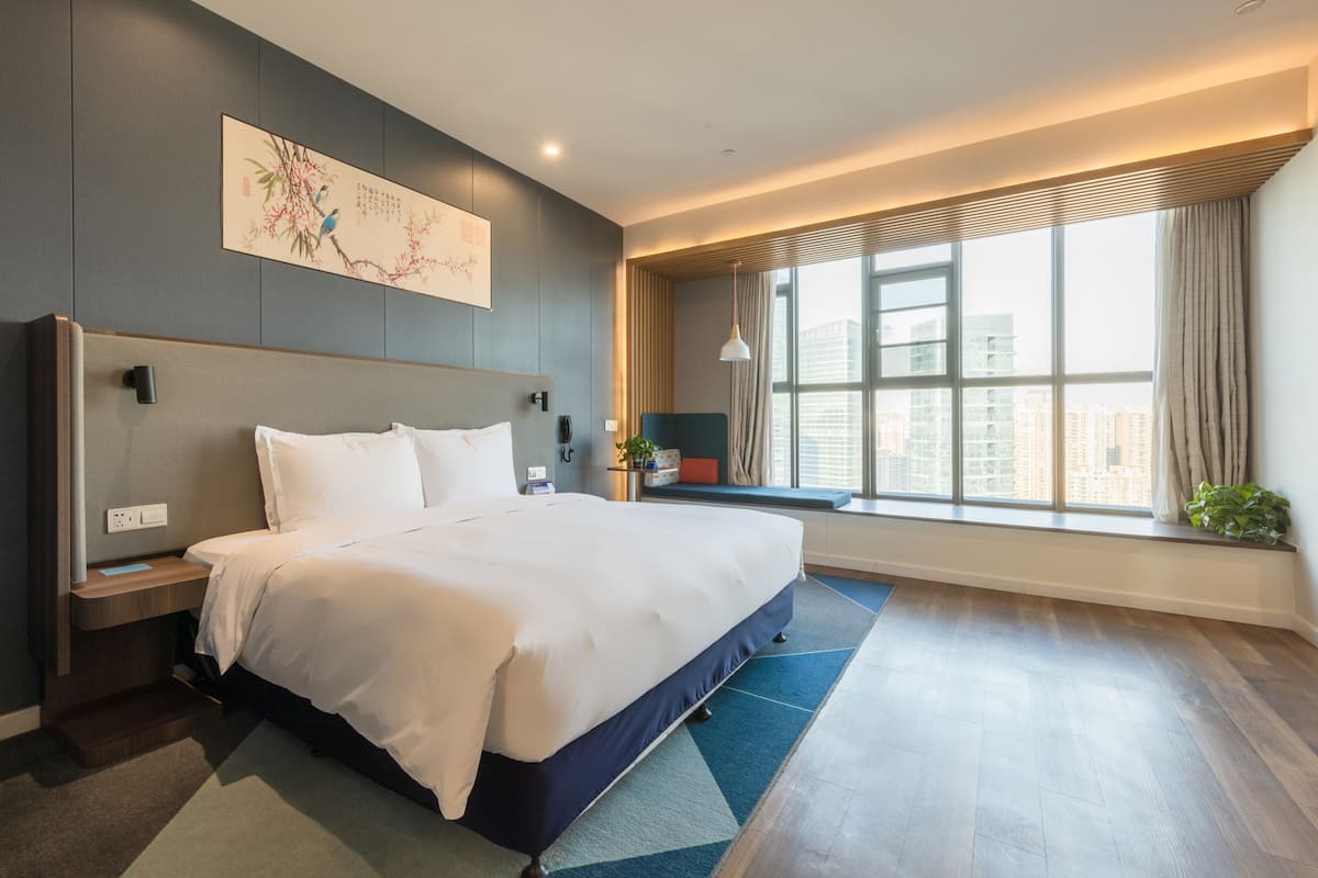 A modern hotel room with a large bed, a nightstand, a wall-mounted telephone, a window with a view of the city, and a decorative painting.