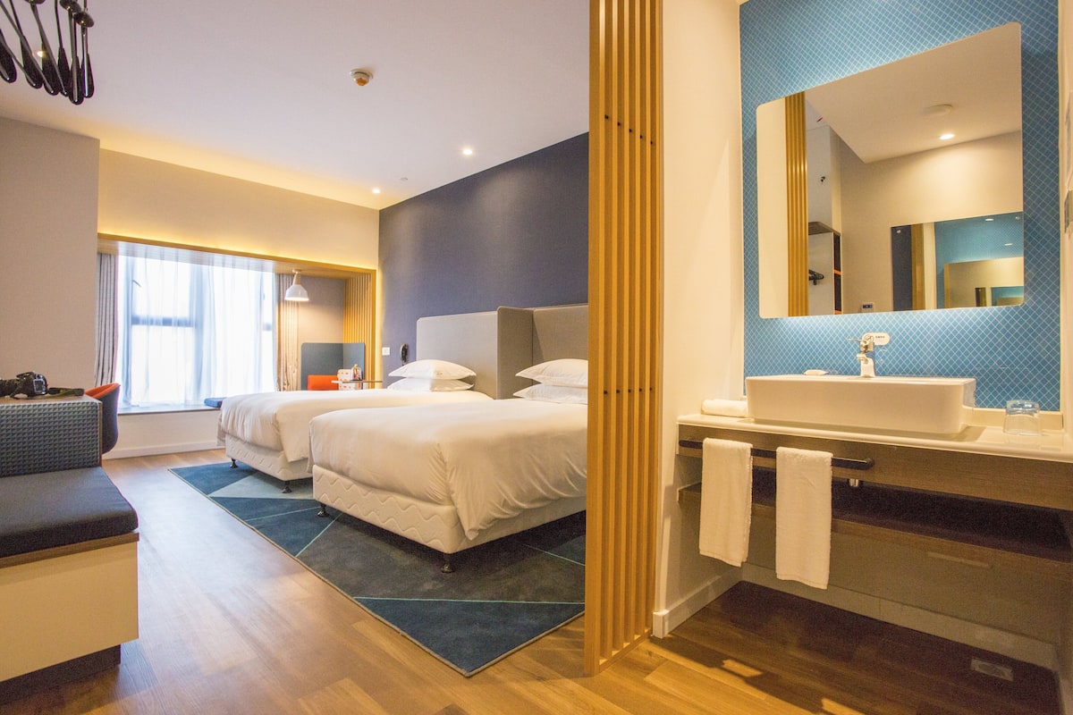 A modern hotel room with a large bed, a separate seating area, a bathroom with a large mirror, and a view of the city.