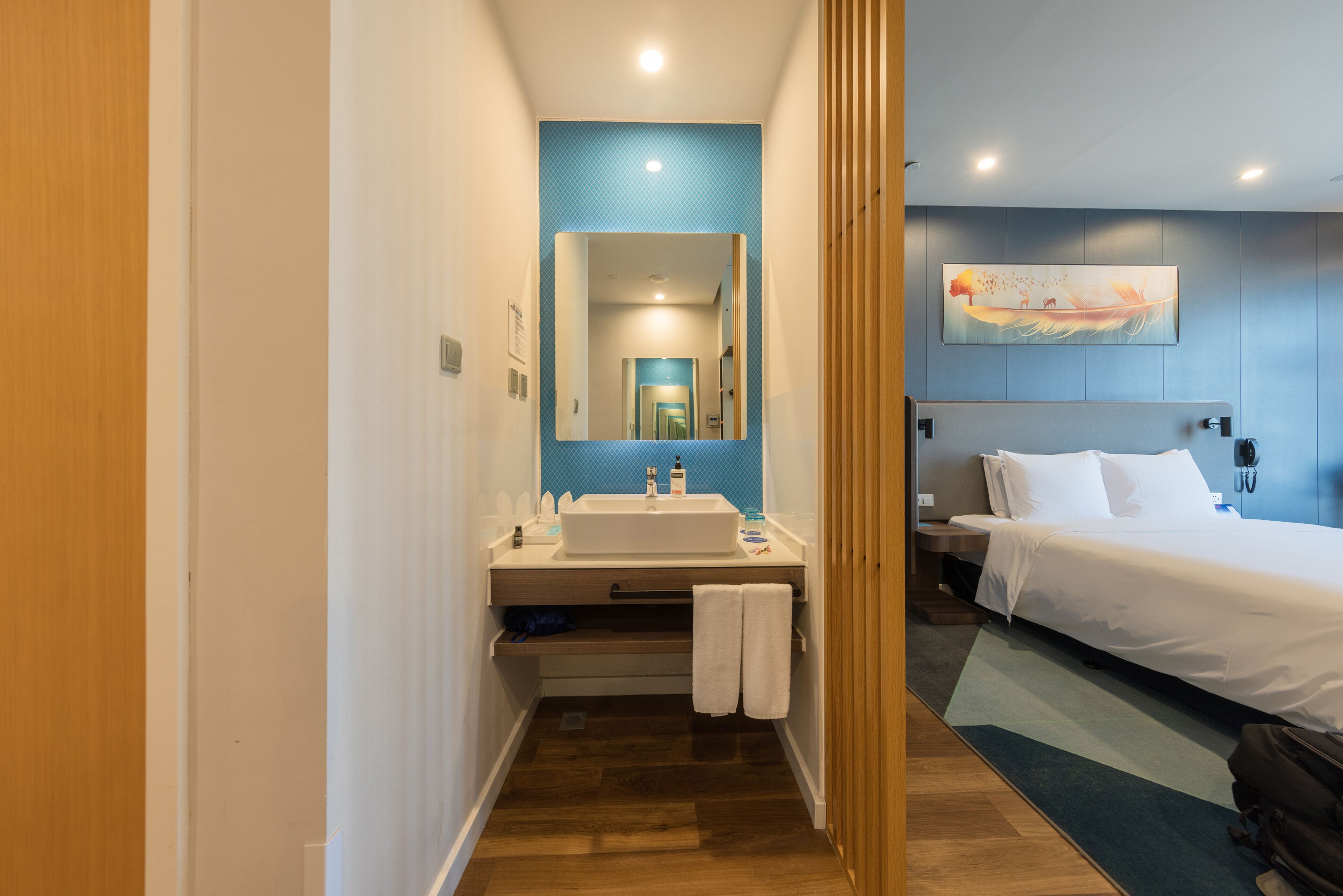 Habitación de hotel con piso de madera, una cama blanca, una mesita de noche, un lavamanos blanco y una pared de acento azul.