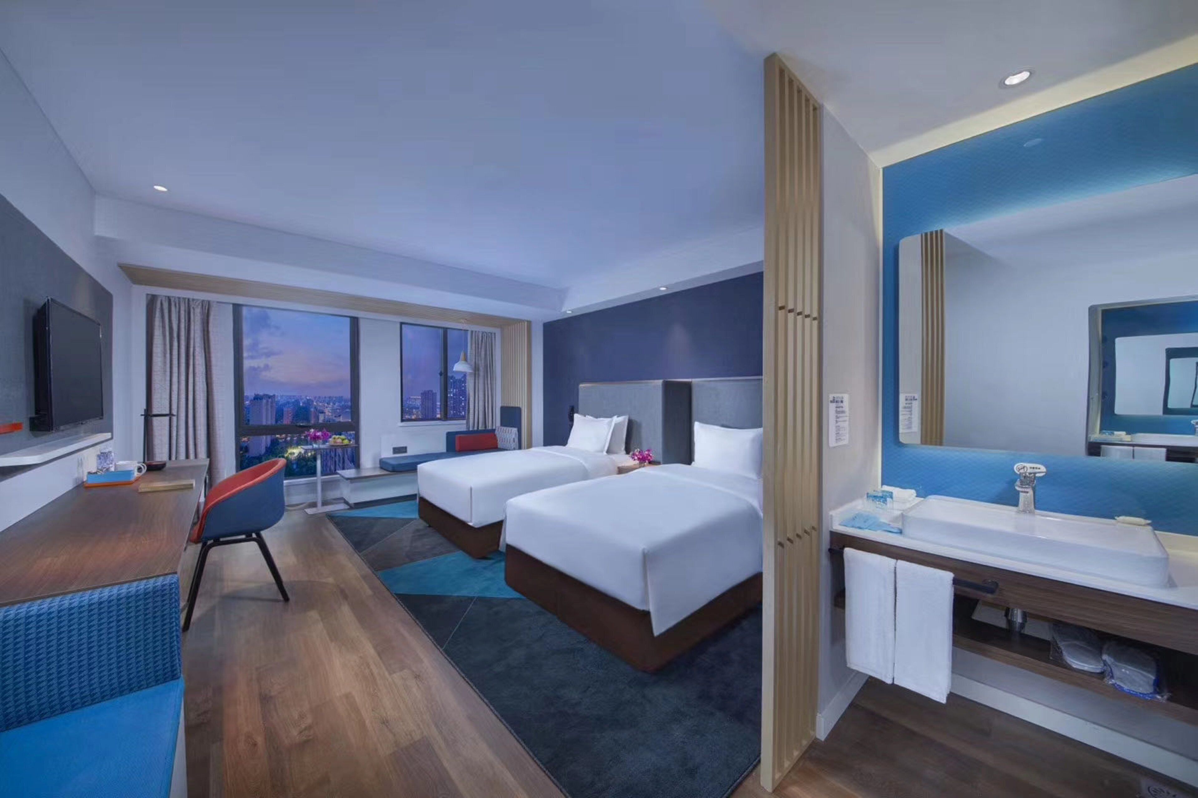 A hotel room with two beds, a desk, a TV, a large window with a city view, and a bathroom with a mirror and sink.