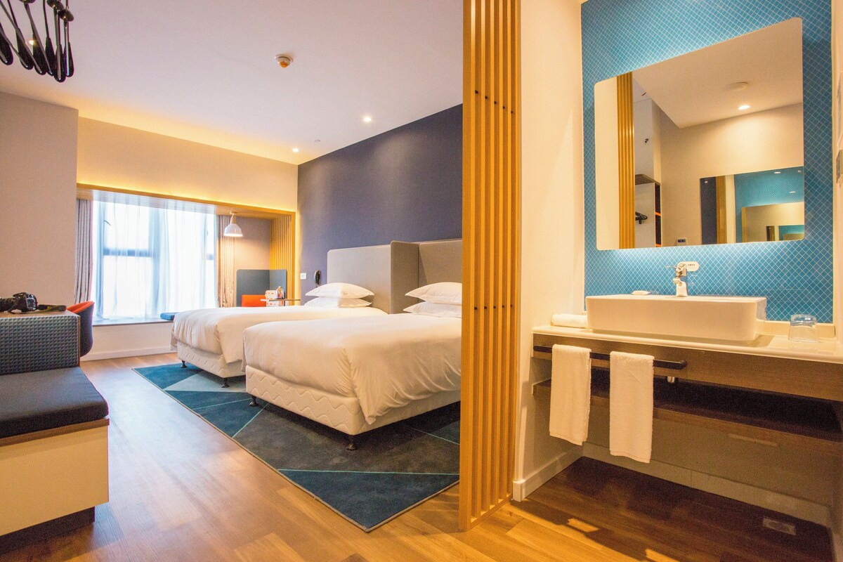 A modern hotel room with a large bed, a separate seating area, a bathroom with a large mirror, and a view of the city.