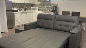 A modern living room with a grey sofa bed, white cabinetry, a kitchen area with a microwave, and a window providing natural light. - Starting Point Family House (Victoria)