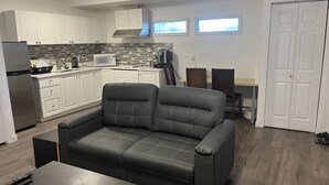 A modern kitchen with white cabinets, a stainless steel refrigerator, and a microwave. There is a gray leather sofa and a wooden dining table with chairs. A clock is visible on the wall. - Starting Point Family House (Victoria)