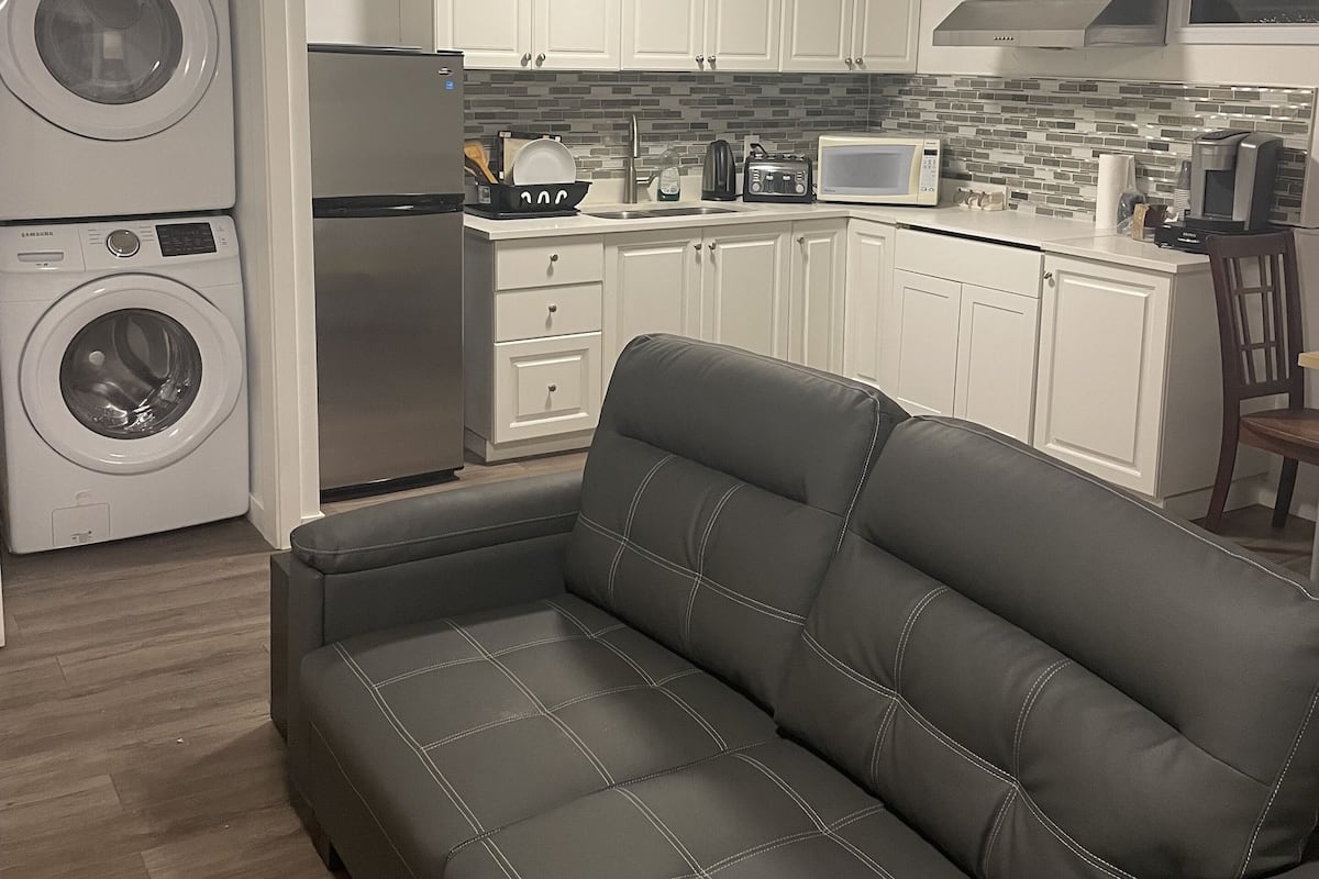 A compact living space with a sofa, washing machine, refrigerator, microwave, and tiled backsplash.