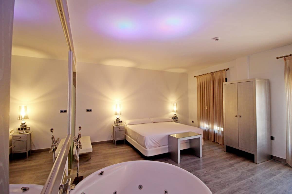 A modern hotel room with a large bed, bedside tables with lamps, a small table, a wardrobe, and a large bathtub.