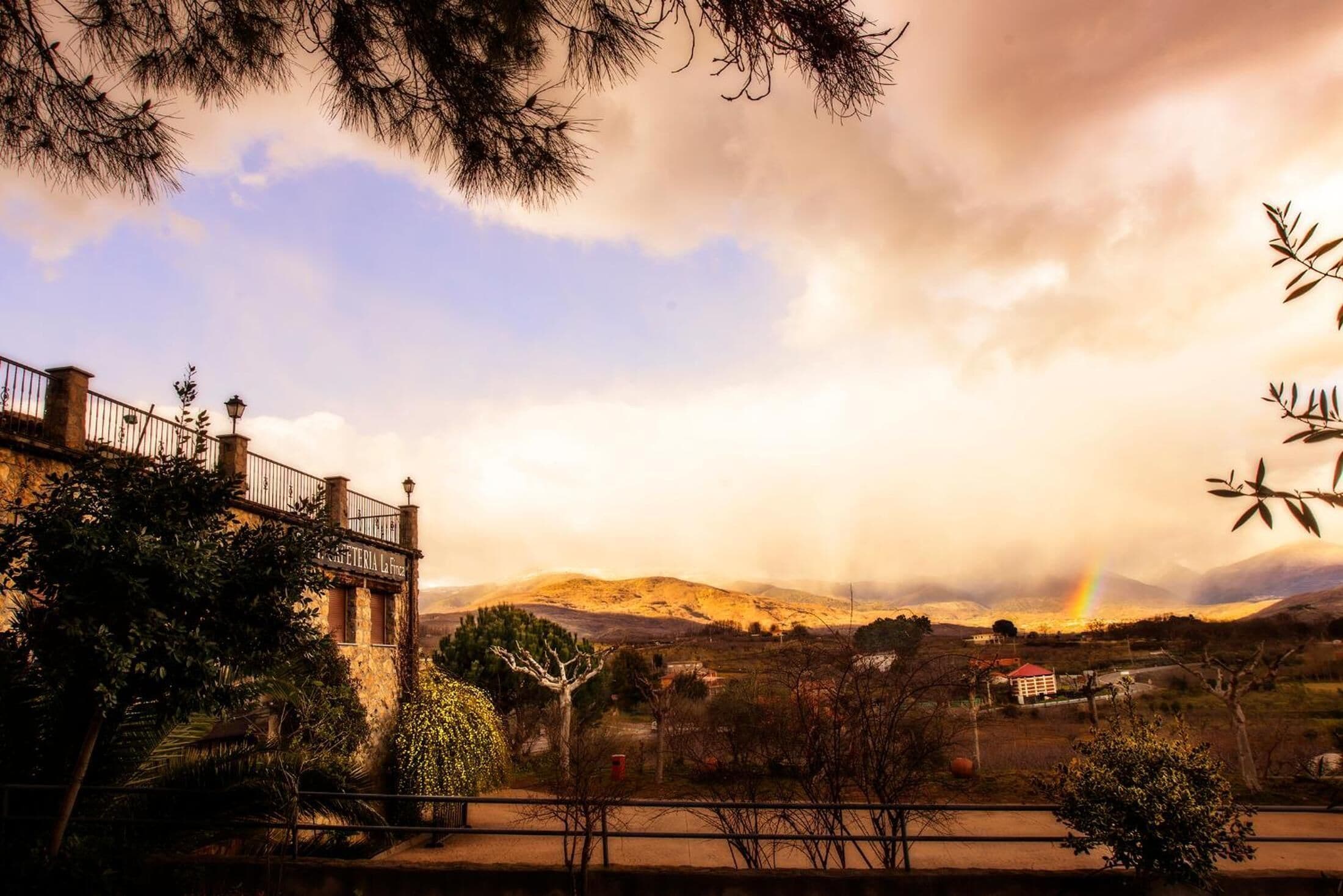 A view of a mountainous landscape with a rainbow, a building with a balcony, and a sign reading "CATERIA La Fiera".