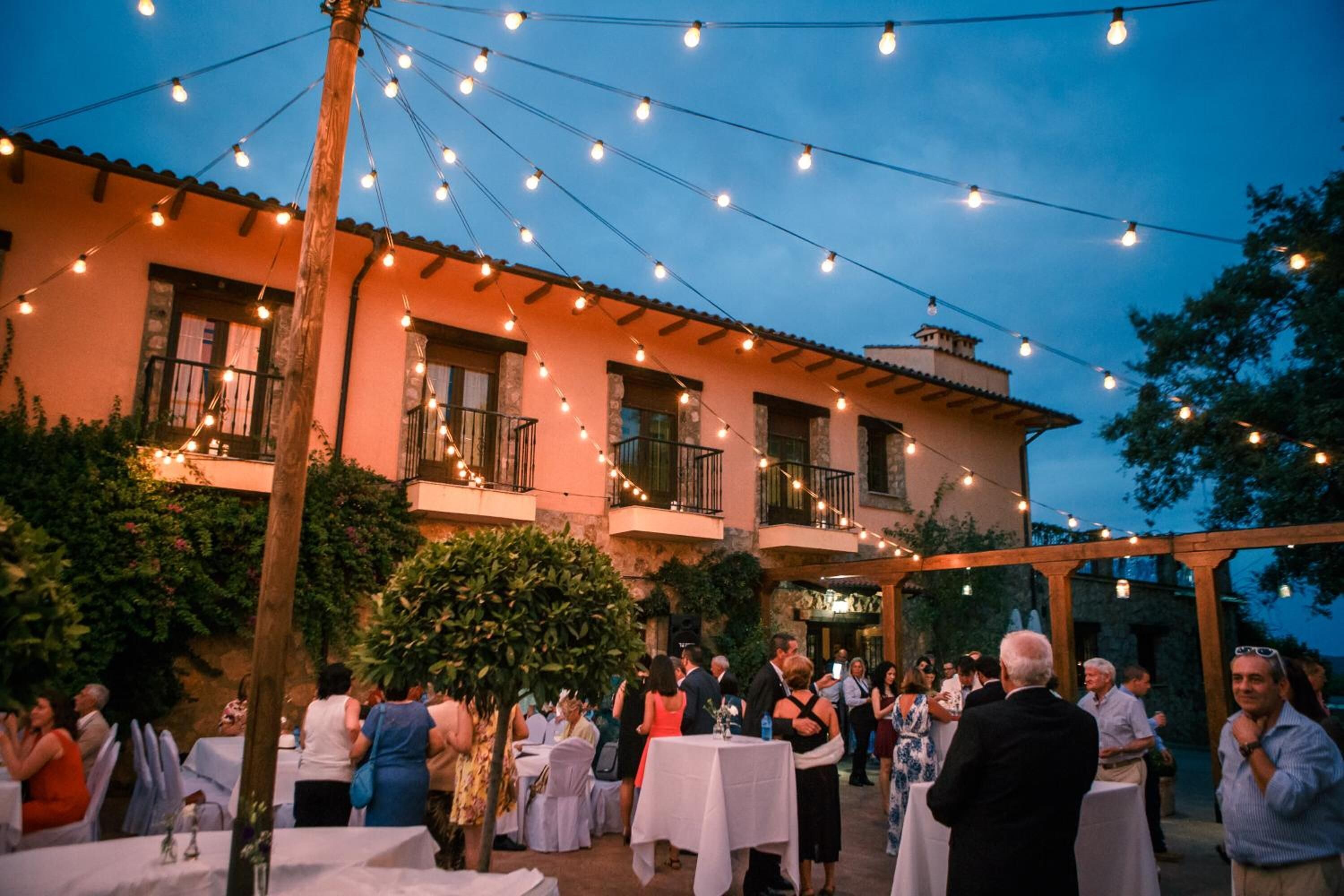 An outdoor event with tables and guests, a building with balconies, and string lights.