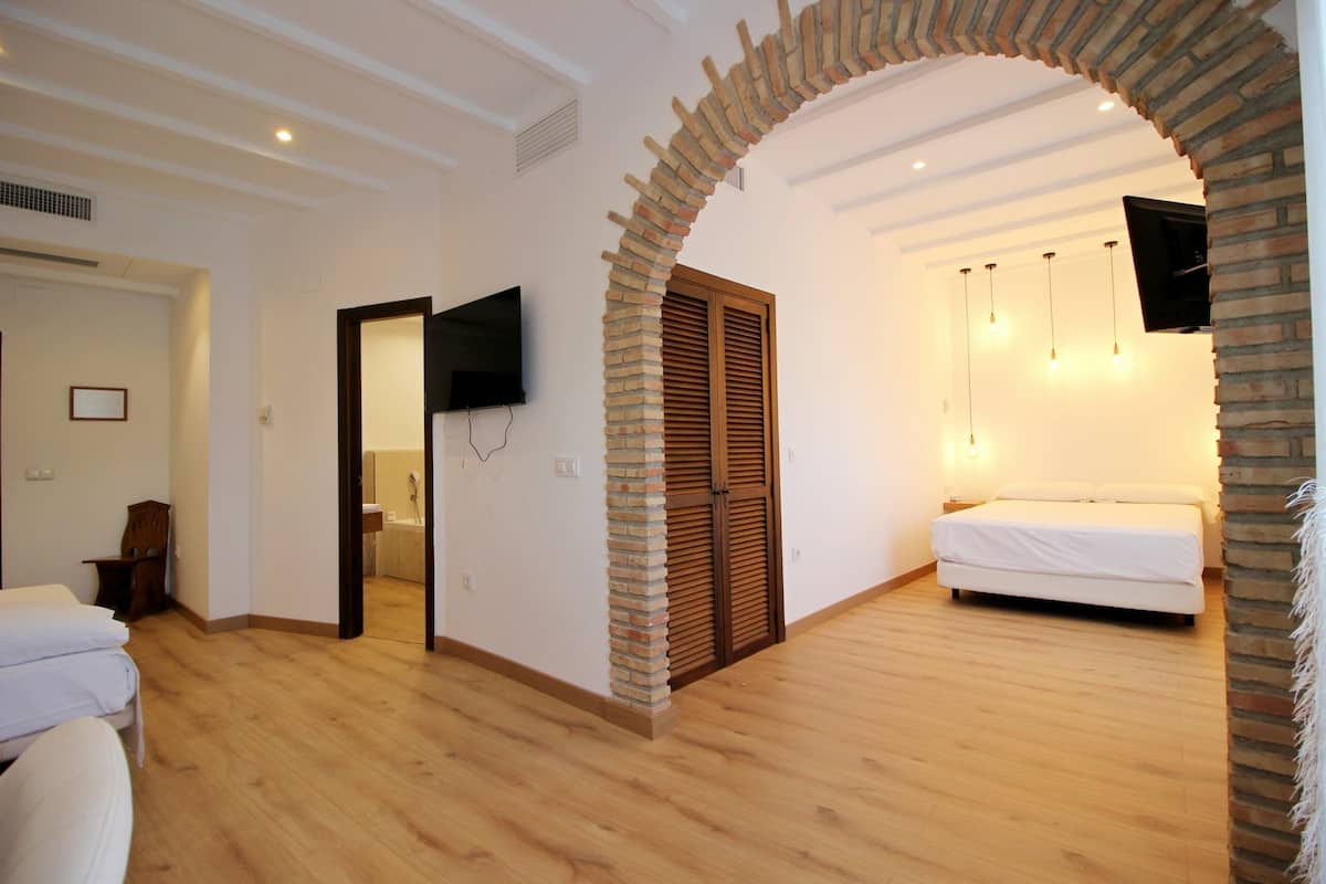 A bedroom with a wooden floor, a bed, a TV, and a brick archway.