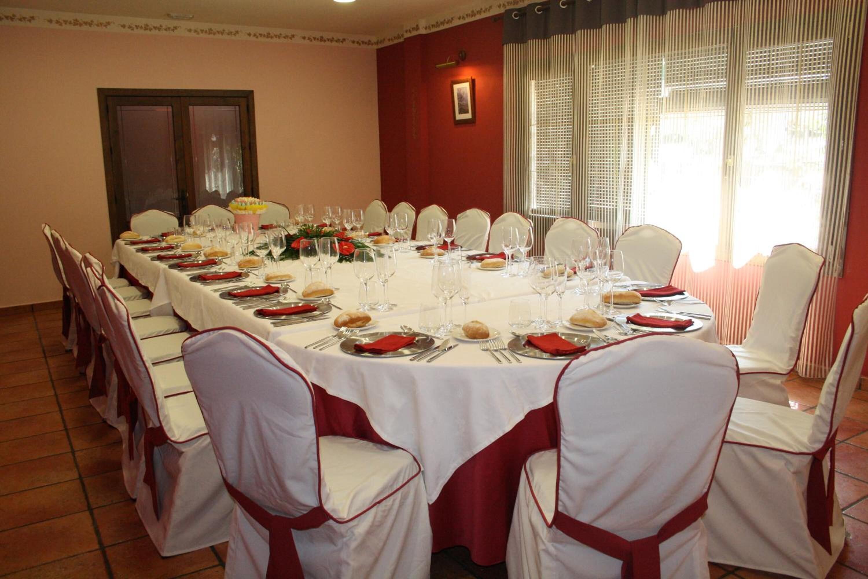 A long dining table set for a formal event, surrounded by white chairs with red sashes, in a room with red walls and a large window.