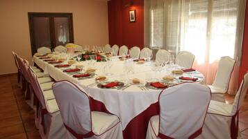 A long dining table set for a formal event, surrounded by white chairs with red sashes, in a room with red walls and a large window.