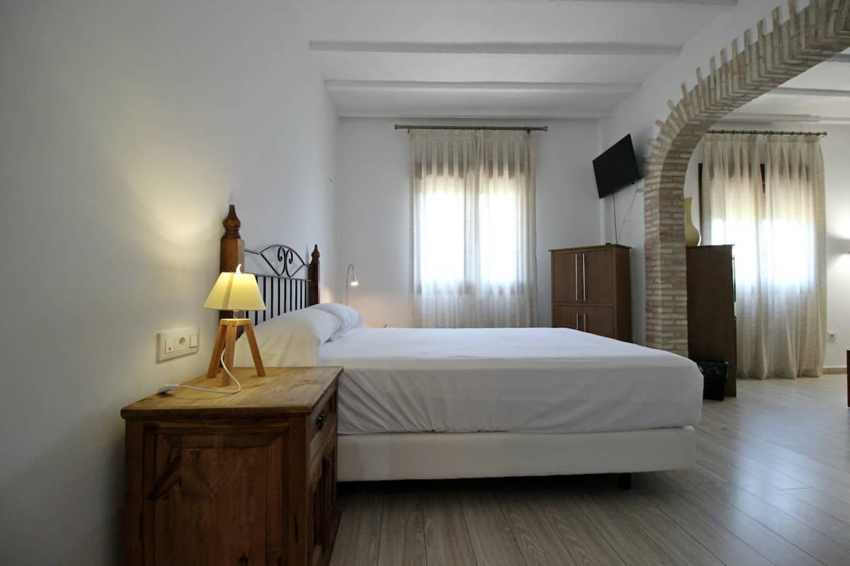 A bedroom with a bed, a wooden nightstand, a lamp, a TV mounted on the wall, and a brick archway.