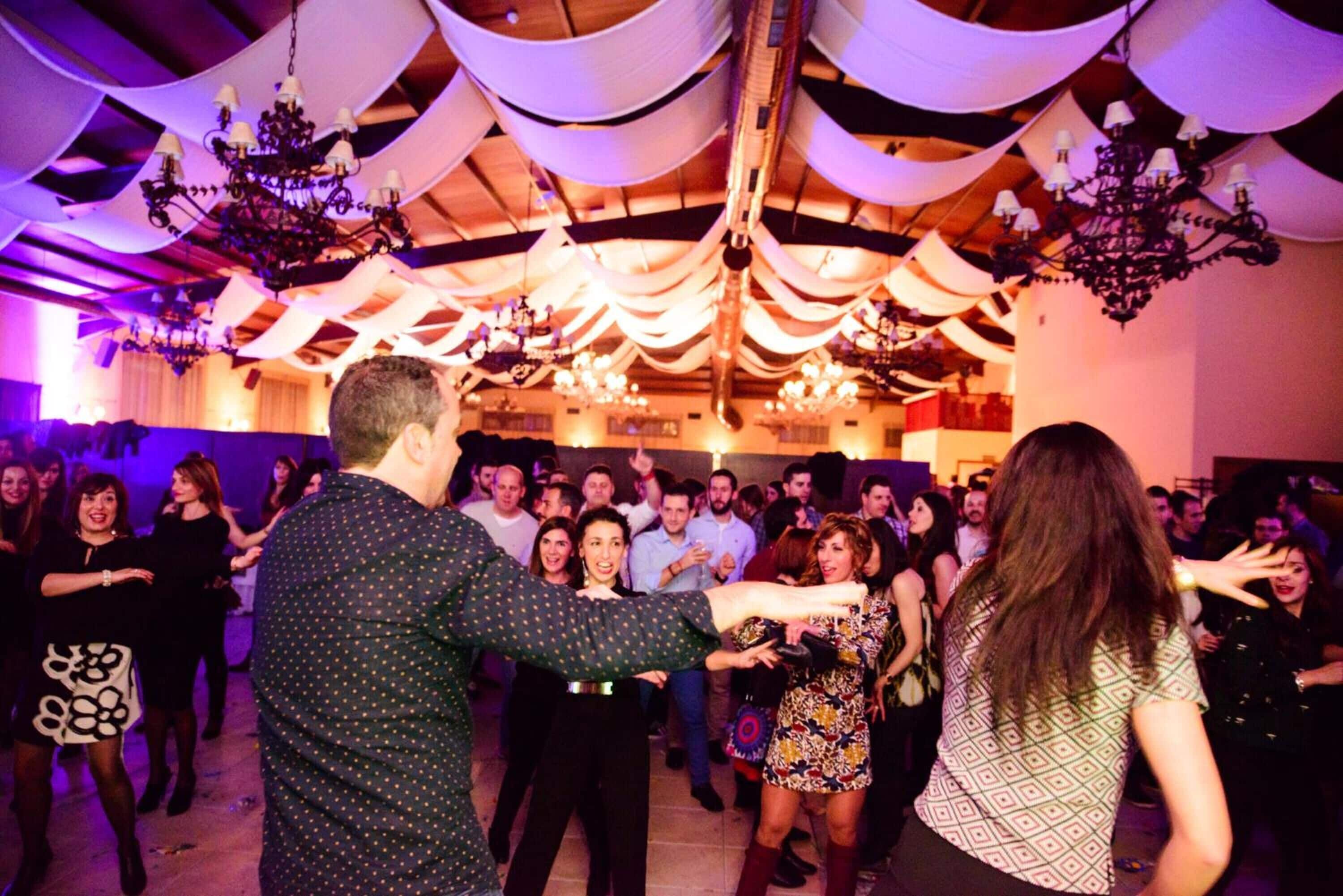 A crowded indoor event with people dancing, chandeliers, and draped fabric decorations.