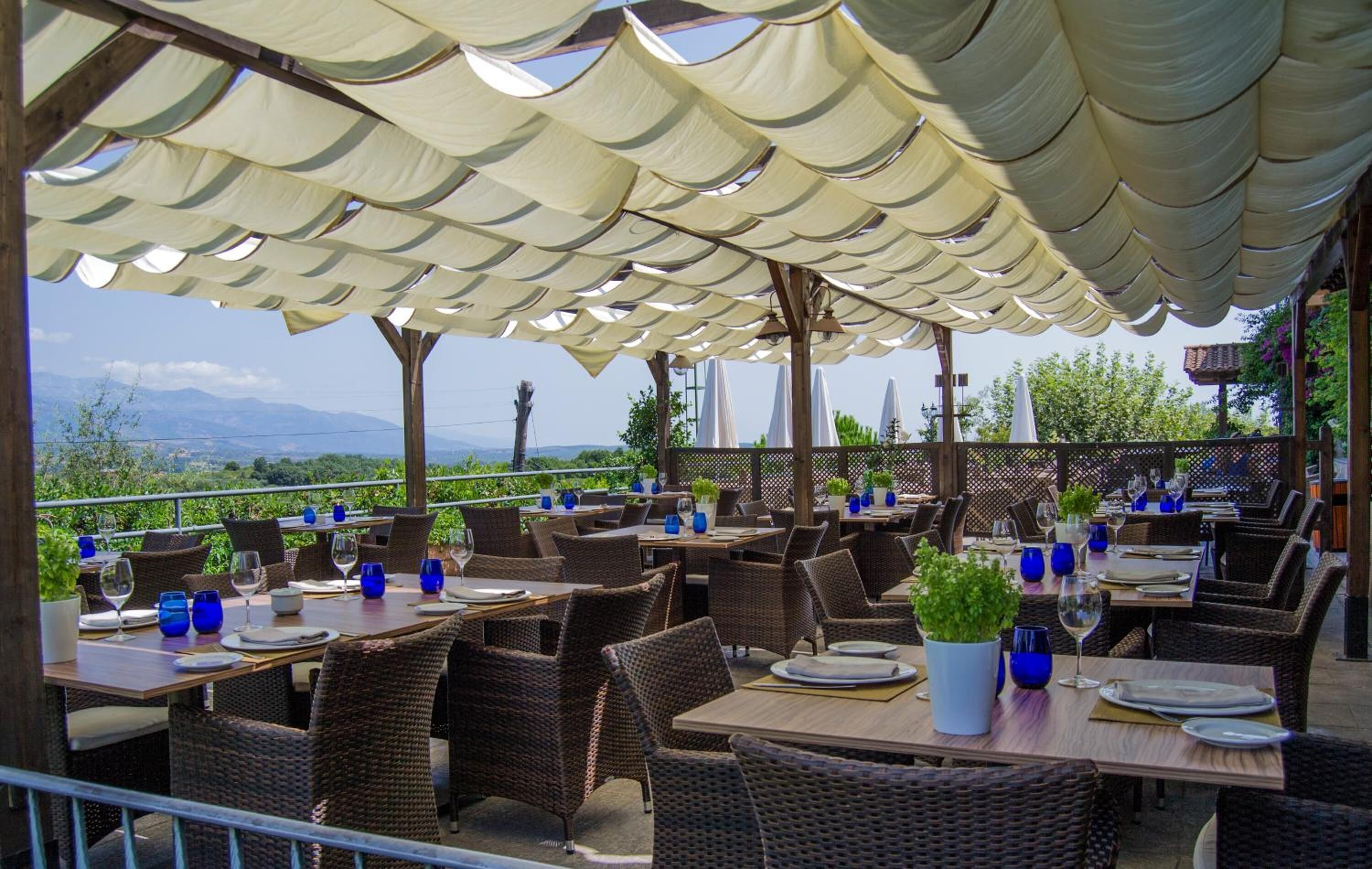An outdoor dining area with wicker chairs and tables, covered by a canopy, and overlooking a scenic landscape.