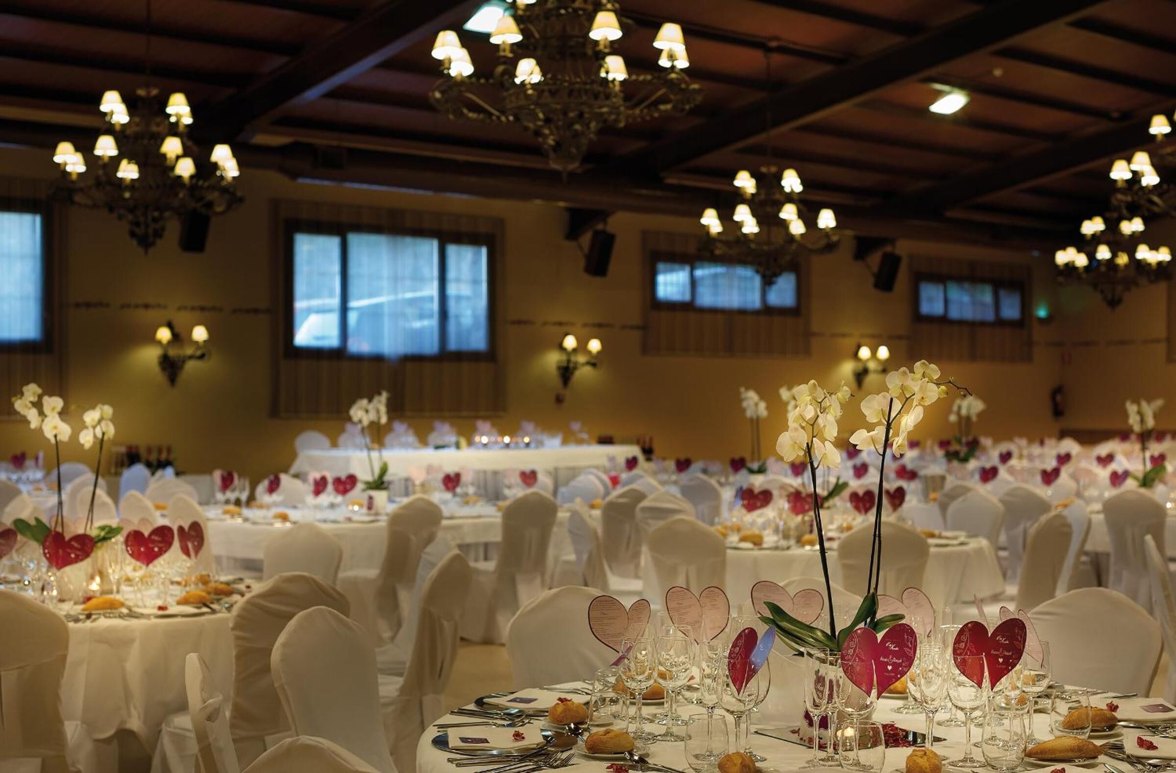 A banquet hall with round tables set for a formal event, featuring heart-shaped centerpieces and white tablecloths.