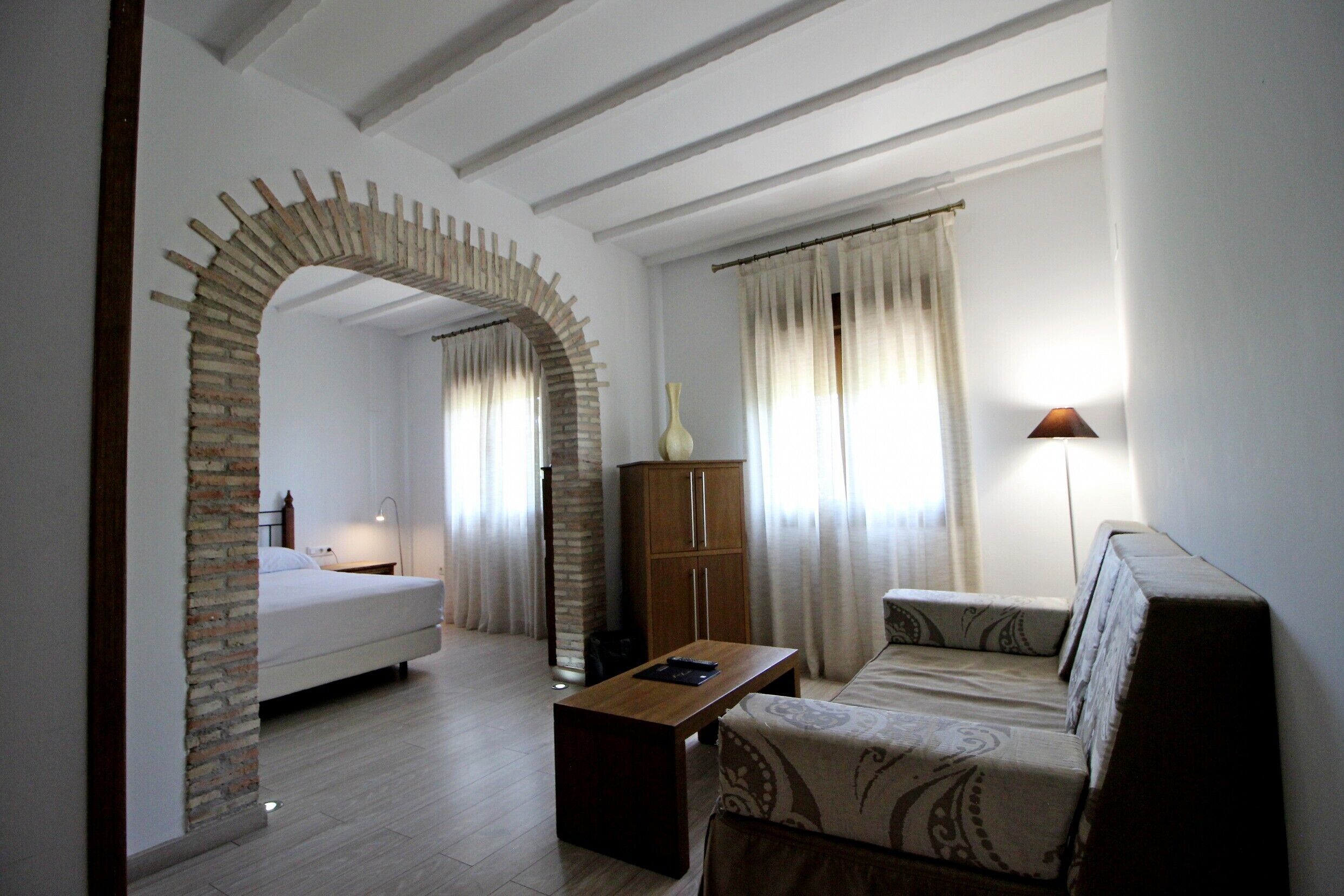 A room with a bed, a sofa, a wooden table, and a lamp. There is a brick archway leading to another room.