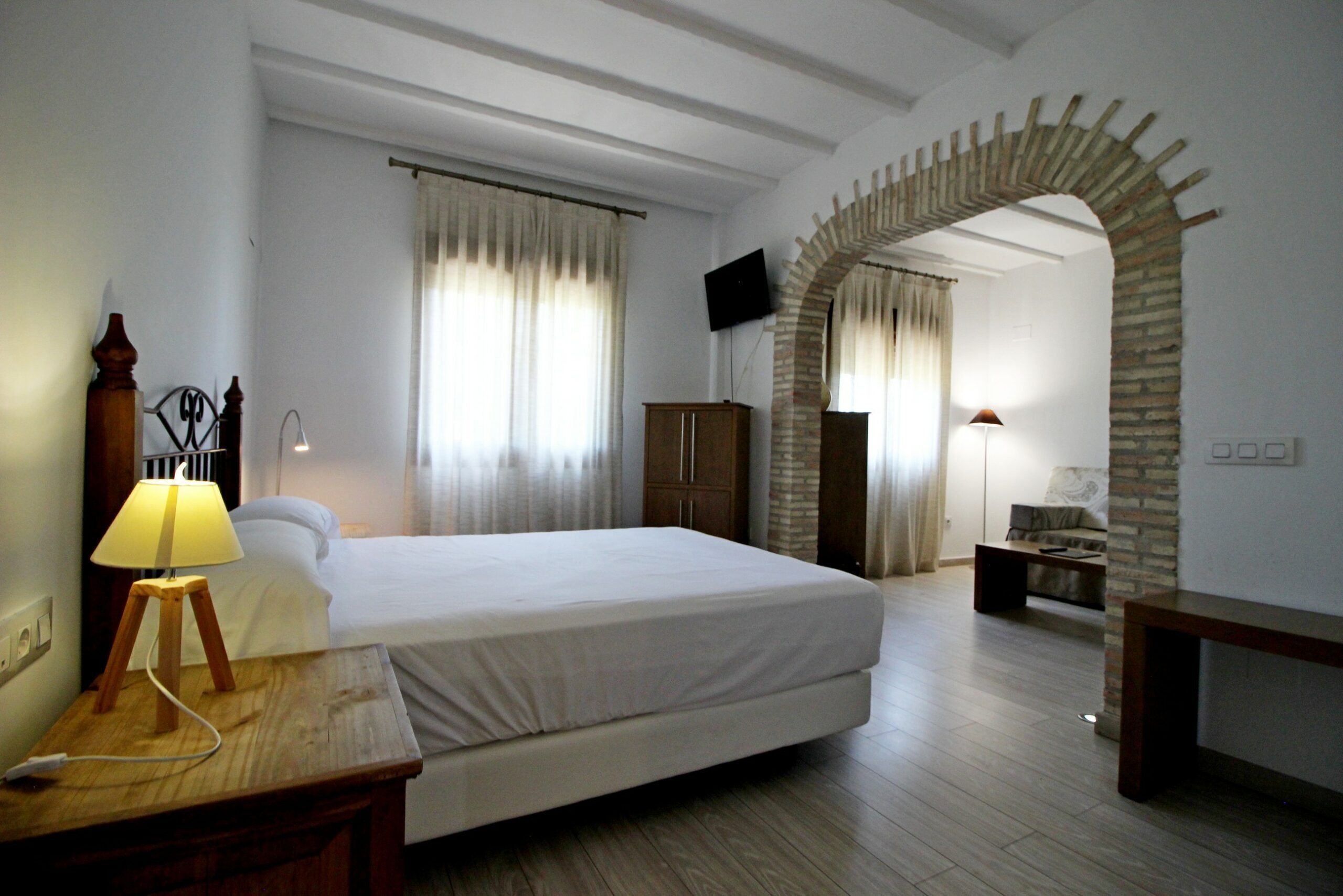 A bedroom with a bed, a wooden nightstand with a lamp, a TV mounted on the wall, and an arched doorway leading to another room.