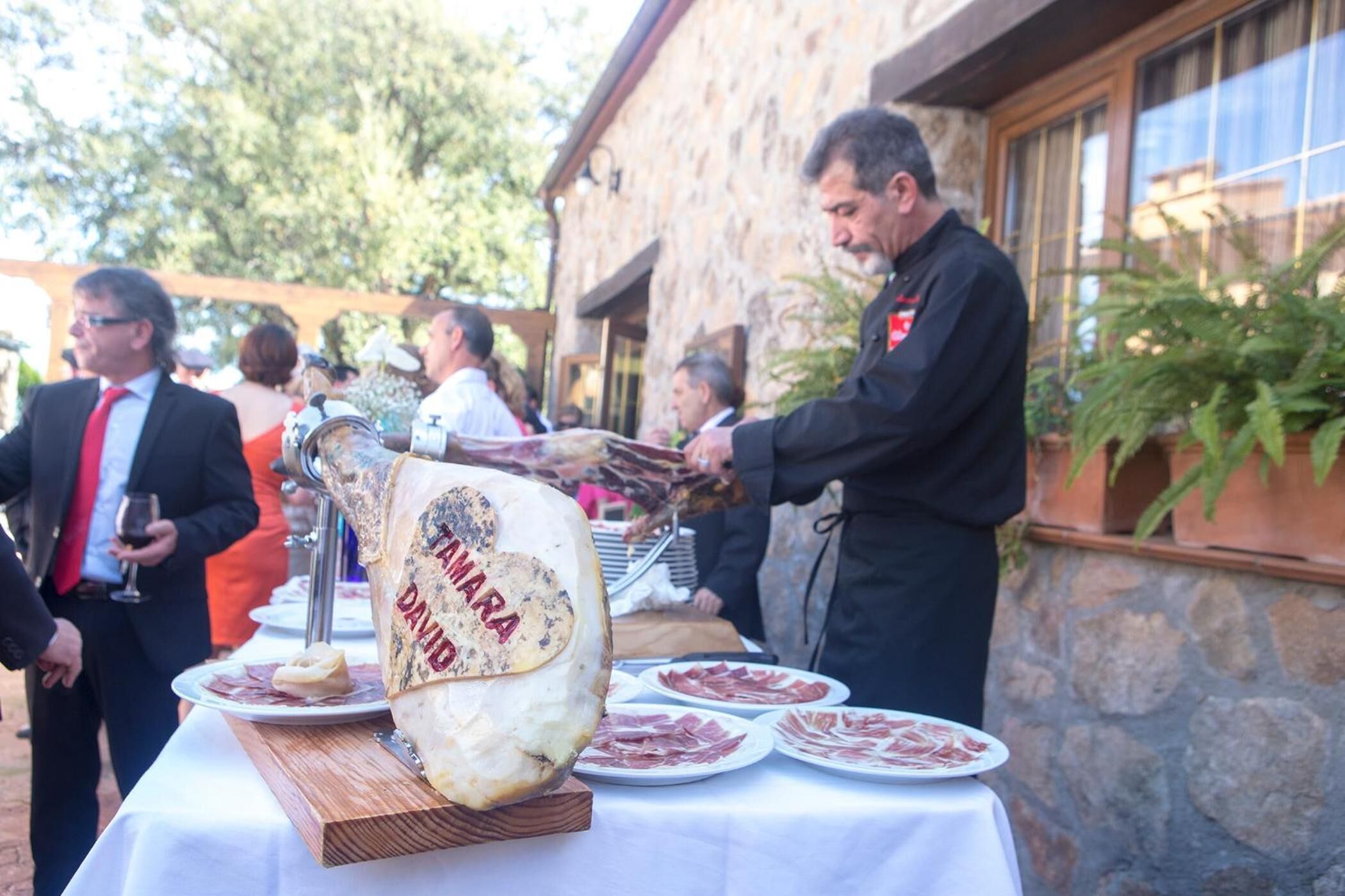 A large wheel of Parmigiano Reggiano cheese on a wooden board, surrounded by plates of cured meats, with people in the background.