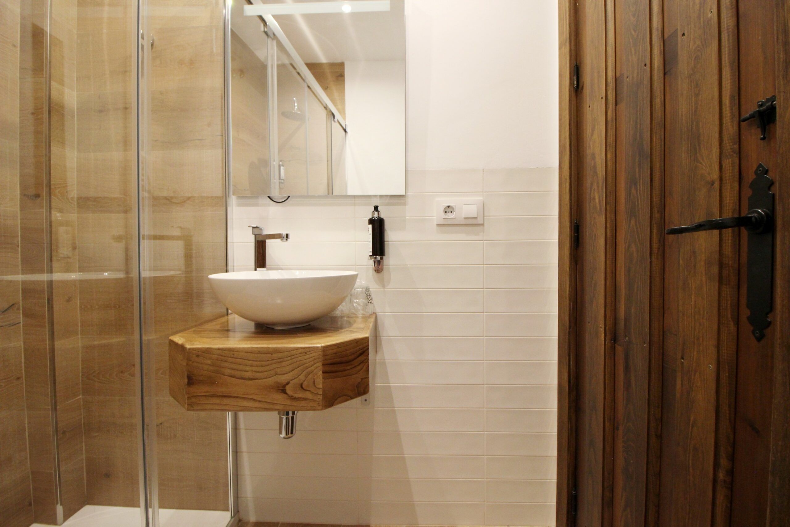 A bathroom with a glass shower enclosure, a wooden vanity with a white sink, and a wooden door.