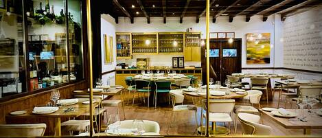 A restaurant with round tables, chairs, and a bar area with shelves of bottles and glasses.