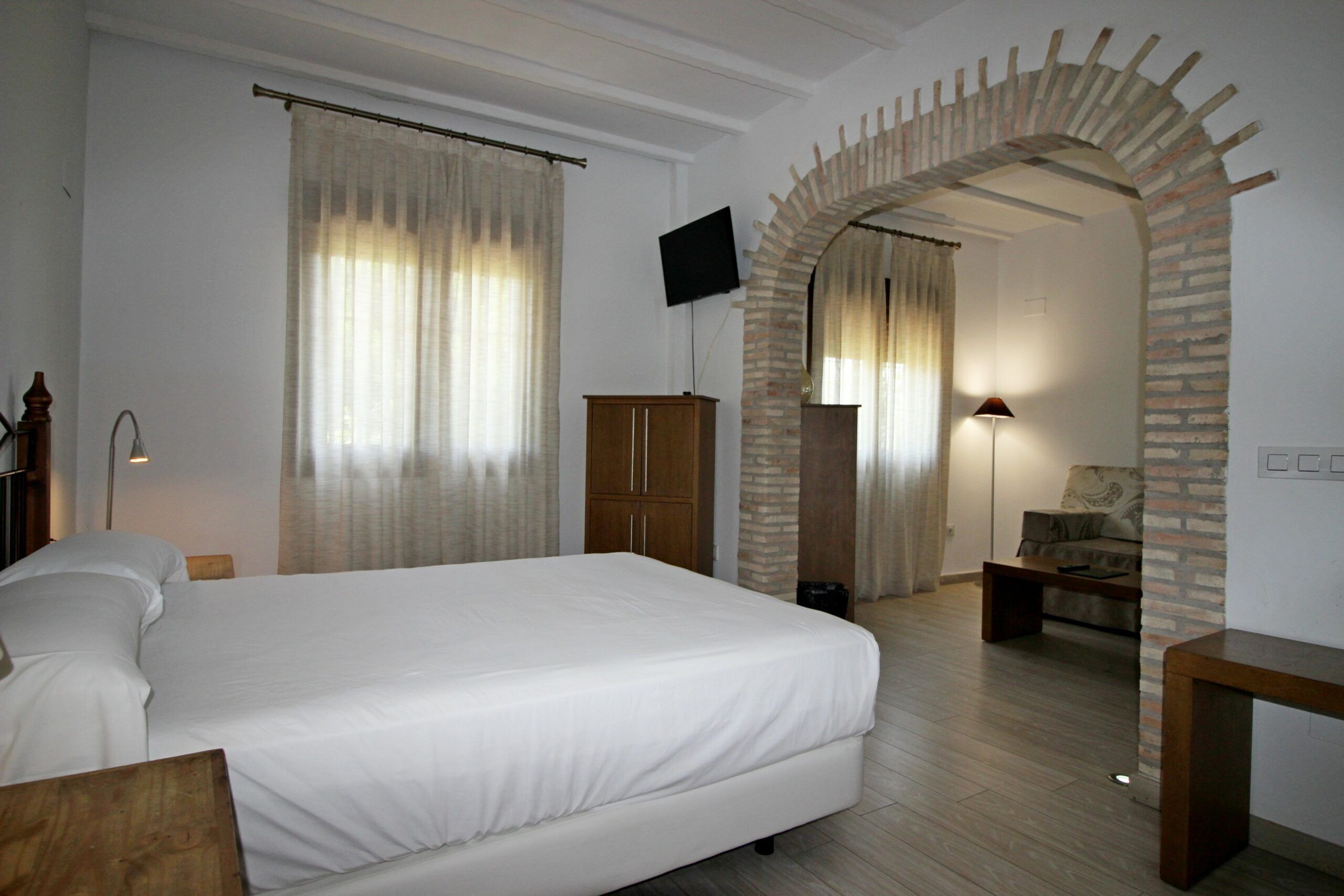 Een slaapkamer met een groot bed, een aan de muur bevestigde televisie, een houten kledingkast en een bakstenen boog die naar een andere kamer leidt.
