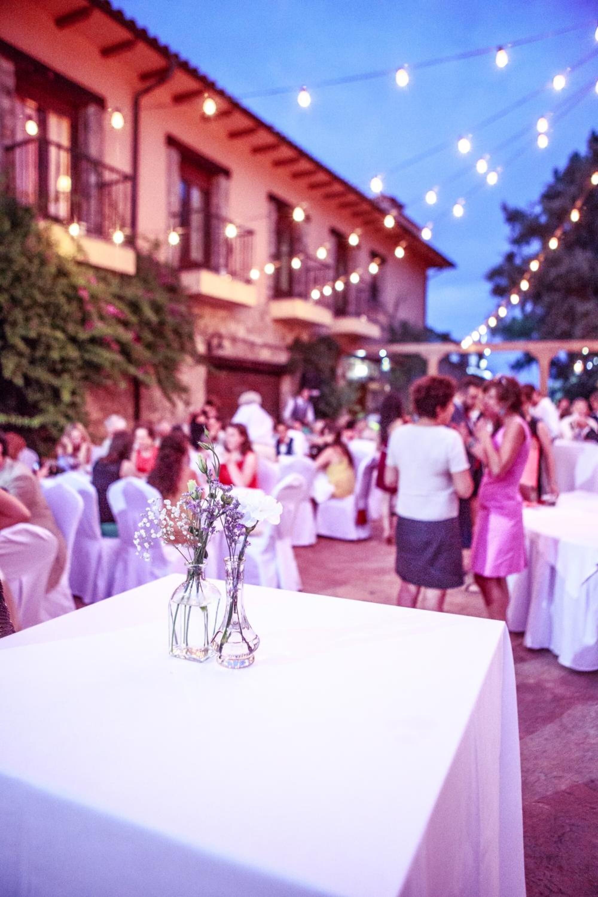 Evento al aire libre con mesas cubiertas de manteles blancos, invitados de pie conversando y luces colgantes.