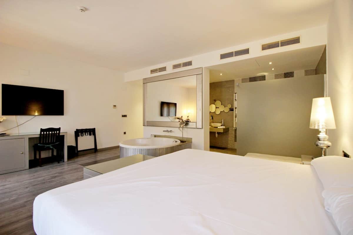 A hotel room with a large bed, a desk with a chair, a TV, a bathtub, and a bathroom with a mirror and sink.