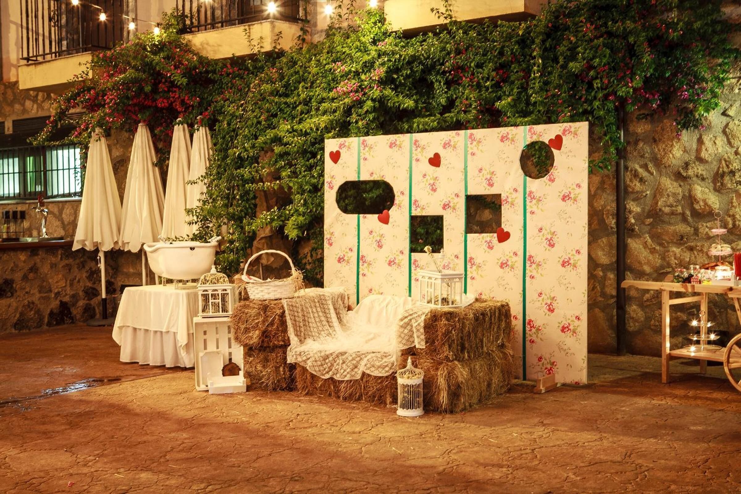 Un entorno rústico al aire libre con fardos de heno, faroles blancos y un decorado con motivos florales y corazones.