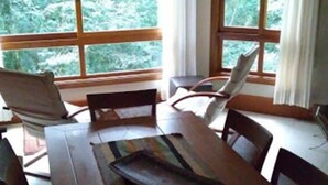 A dining area with a wooden table and chairs, large windows with curtains, and a view of greenery outside. - Locar-in Gramado Vista verde e perto de Tudo (Gramado)