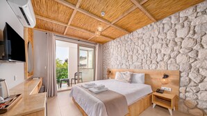 Una habitación de hotel moderna con pared de piedra, techo de madera, cama con sábanas blancas, mesita de noche con lámpara y televisor montado en la pared.