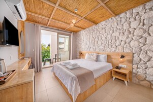 Una habitación de hotel moderna con pared de piedra, techo de madera, cama con sábanas blancas, mesita de noche con lámpara y televisor montado en la pared.