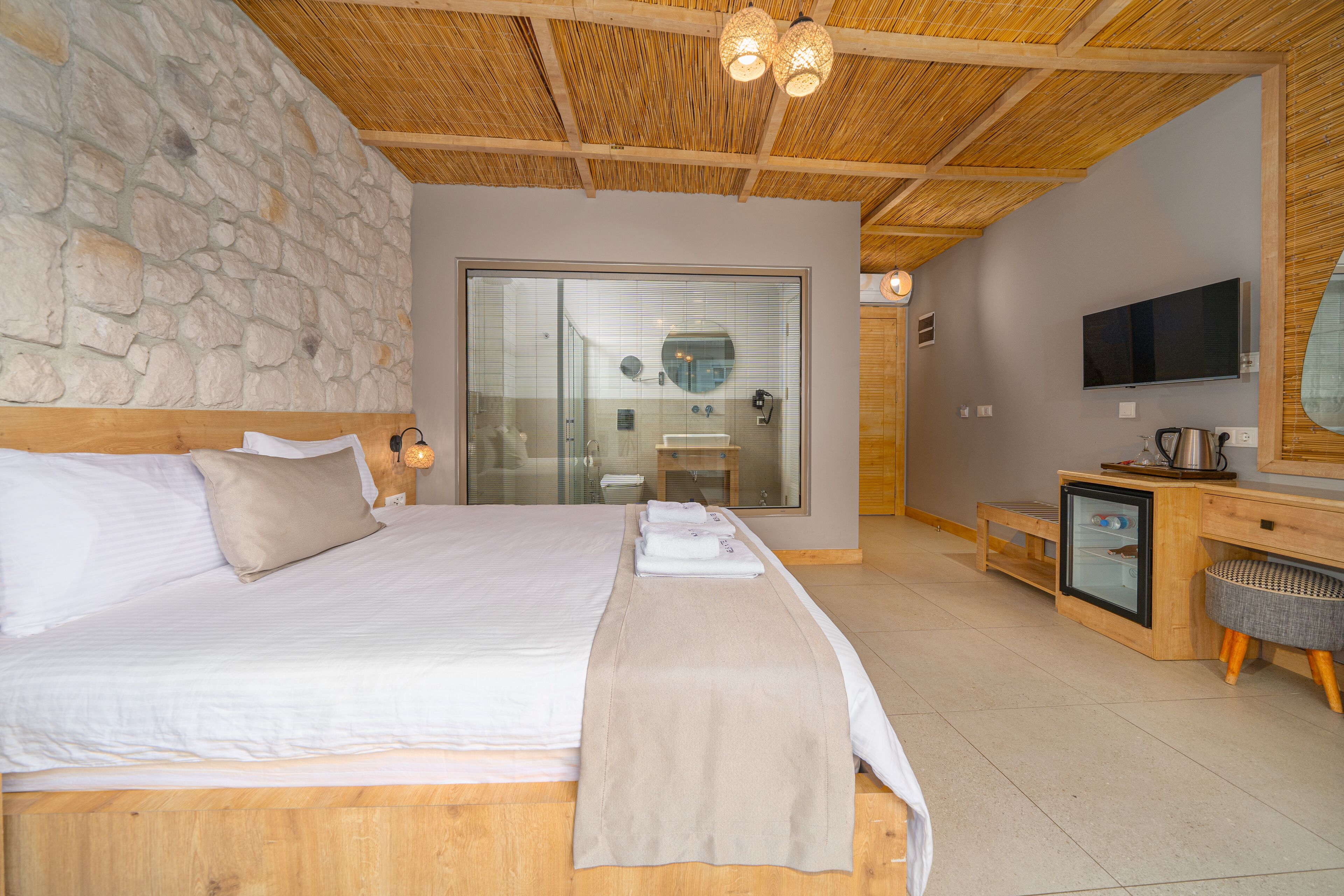 Une chambre avec un grand lit, une tête de lit en bois, un mur en pierre, une porte en verre donnant sur une salle de bain, un bureau en bois avec une télévision et un petit réfrigérateur.