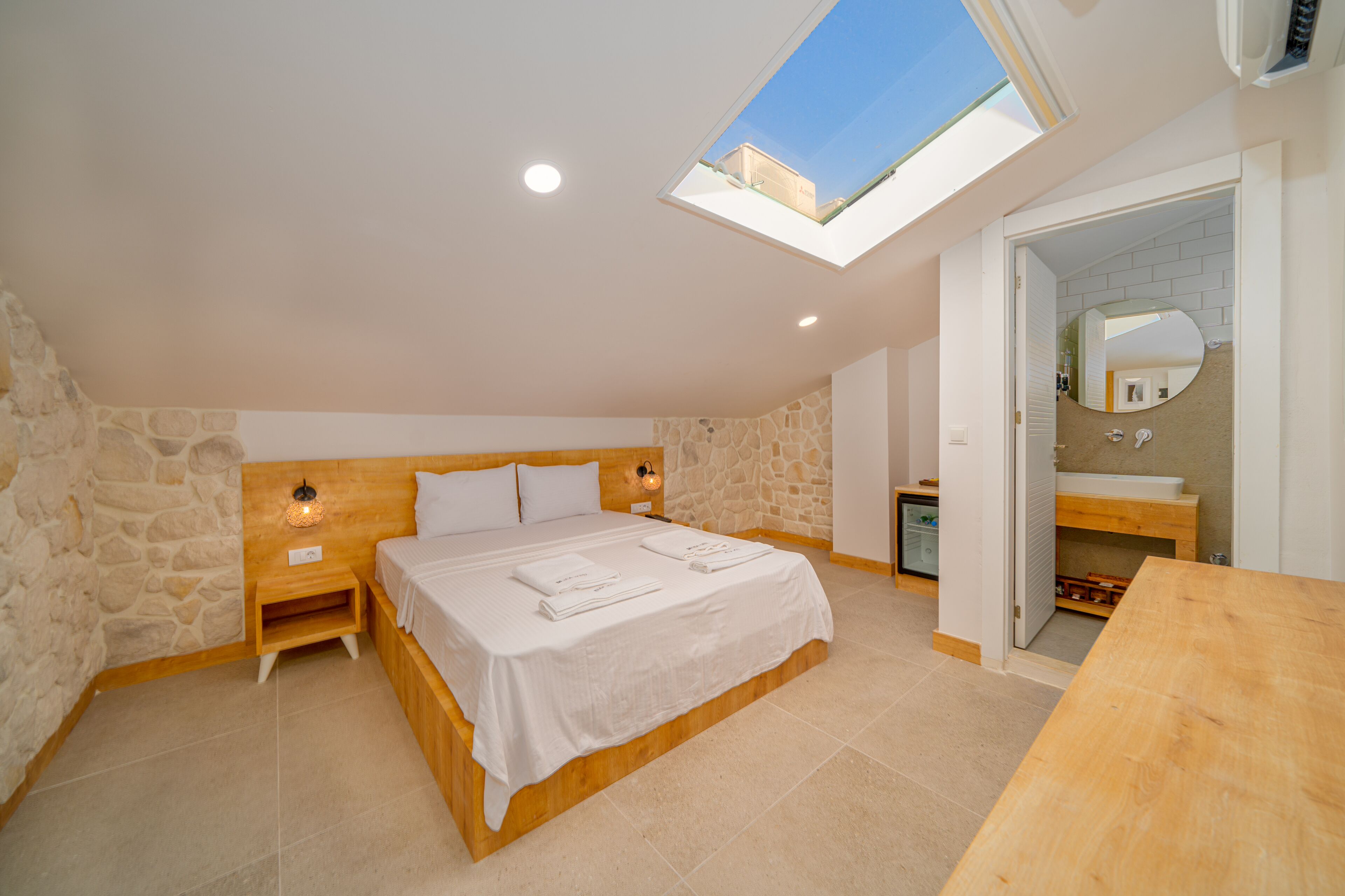 Une chambre avec une lucarne, un lit recouvert de draps blancs, une table de chevet en bois et un mur en pierre.