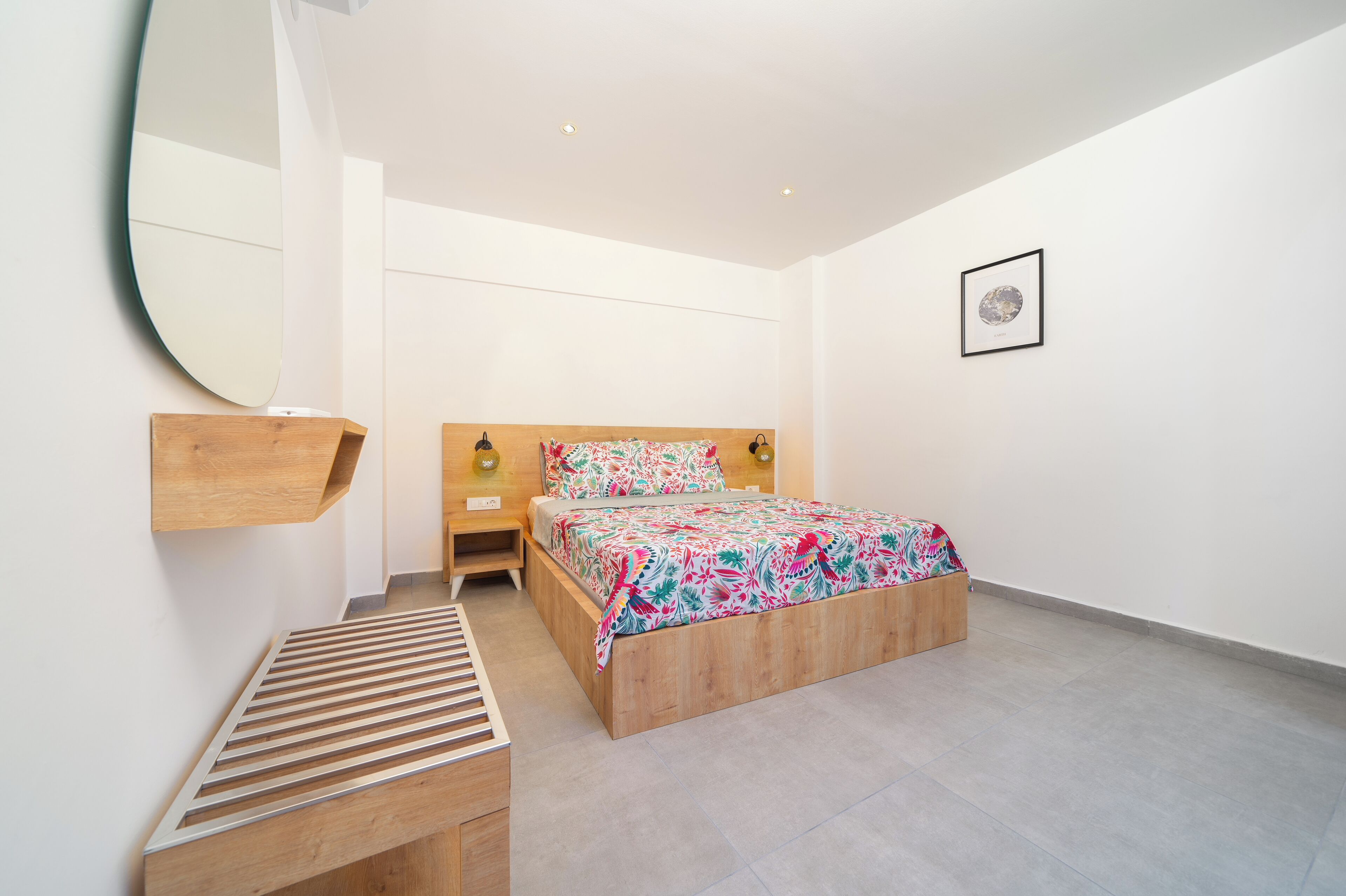 Un dormitorio con una cama, un banco de madera, una mesita de noche, un espejo montado en la pared y un cuadro enmarcado.