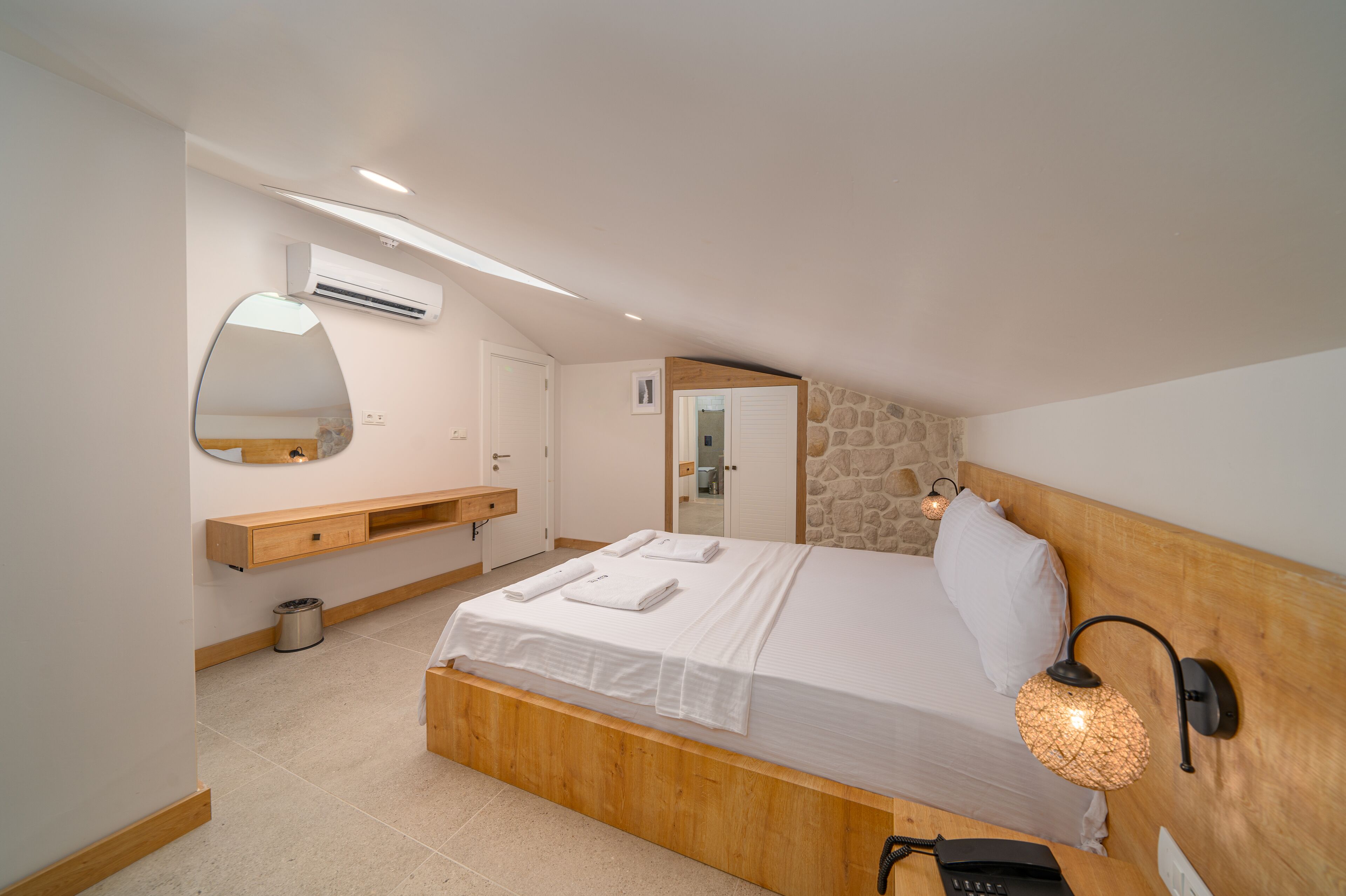 A bedroom with a wooden bed, white linens, a mirror, an air conditioning unit, a stone wall, and a nightstand with a lamp.